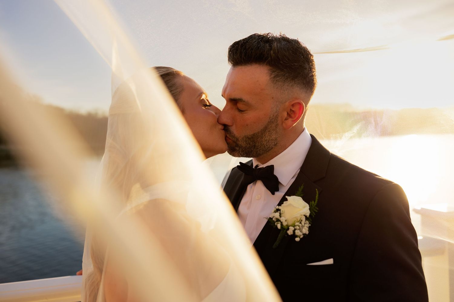 Bride and groom share a romantic kiss outdoors at sunset, with the bride's veil flowing in the breeze.