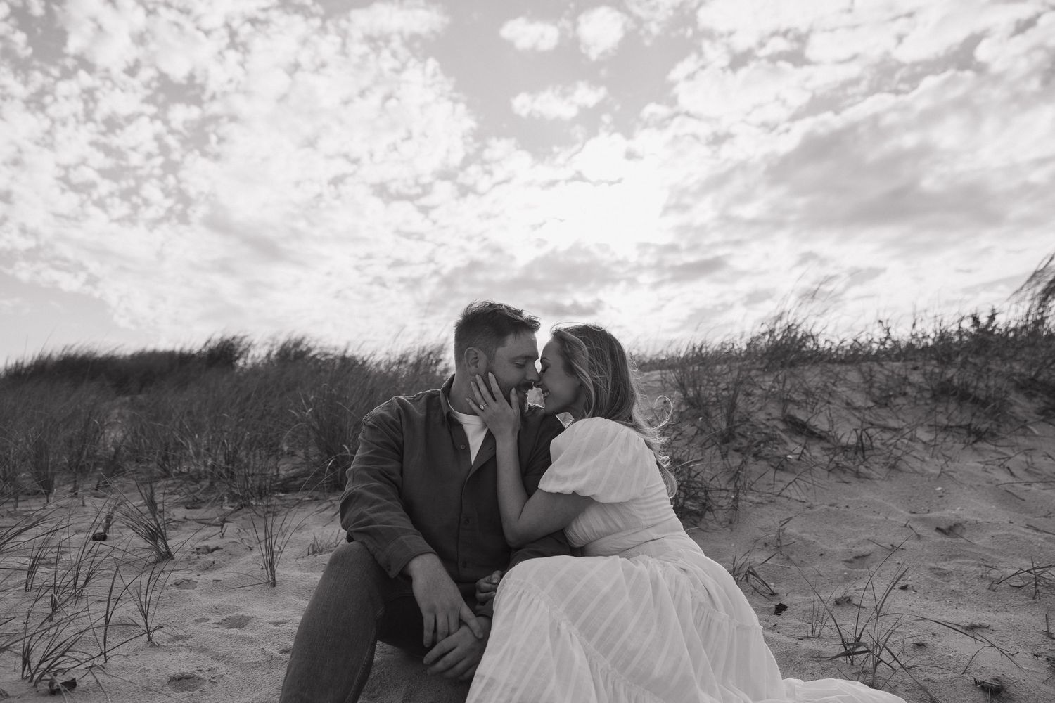 A romantic black and white photograph of a couple sitting close together on sand dunes under a cloudy sky.