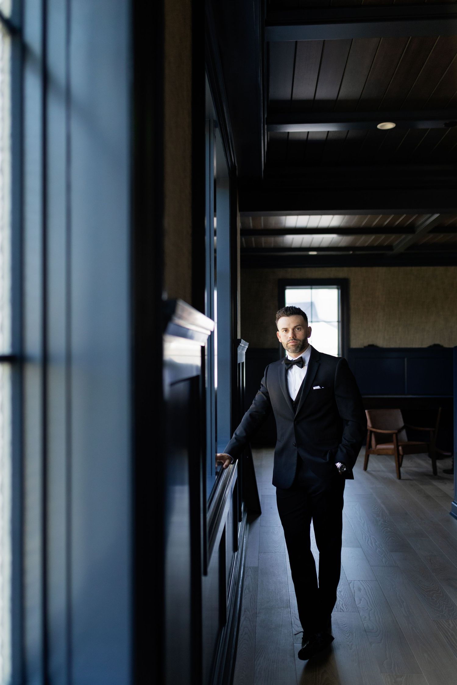 Elegant groom in black tuxedo and bow tie standing in a dark wood-paneled hallway with natural light.