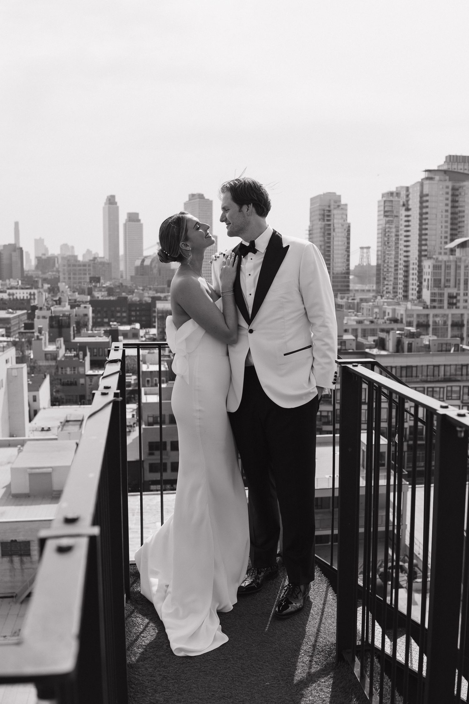 A romantic wedding couple embrace on a rooftop balcony overlooking a city skyline on a cloudy afternoon.