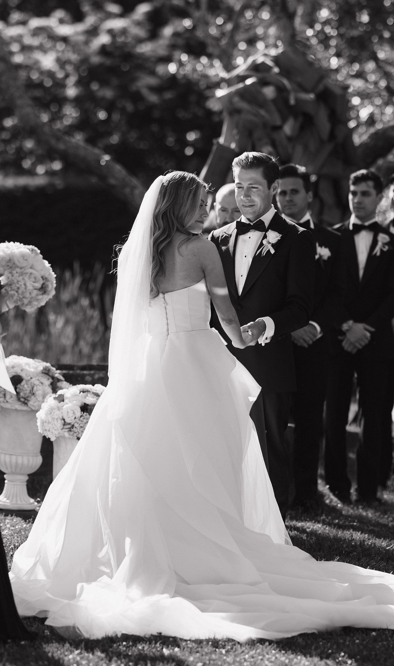 Wedding party gathers outdoors with flowing white gown and formal attire in black and white photography.