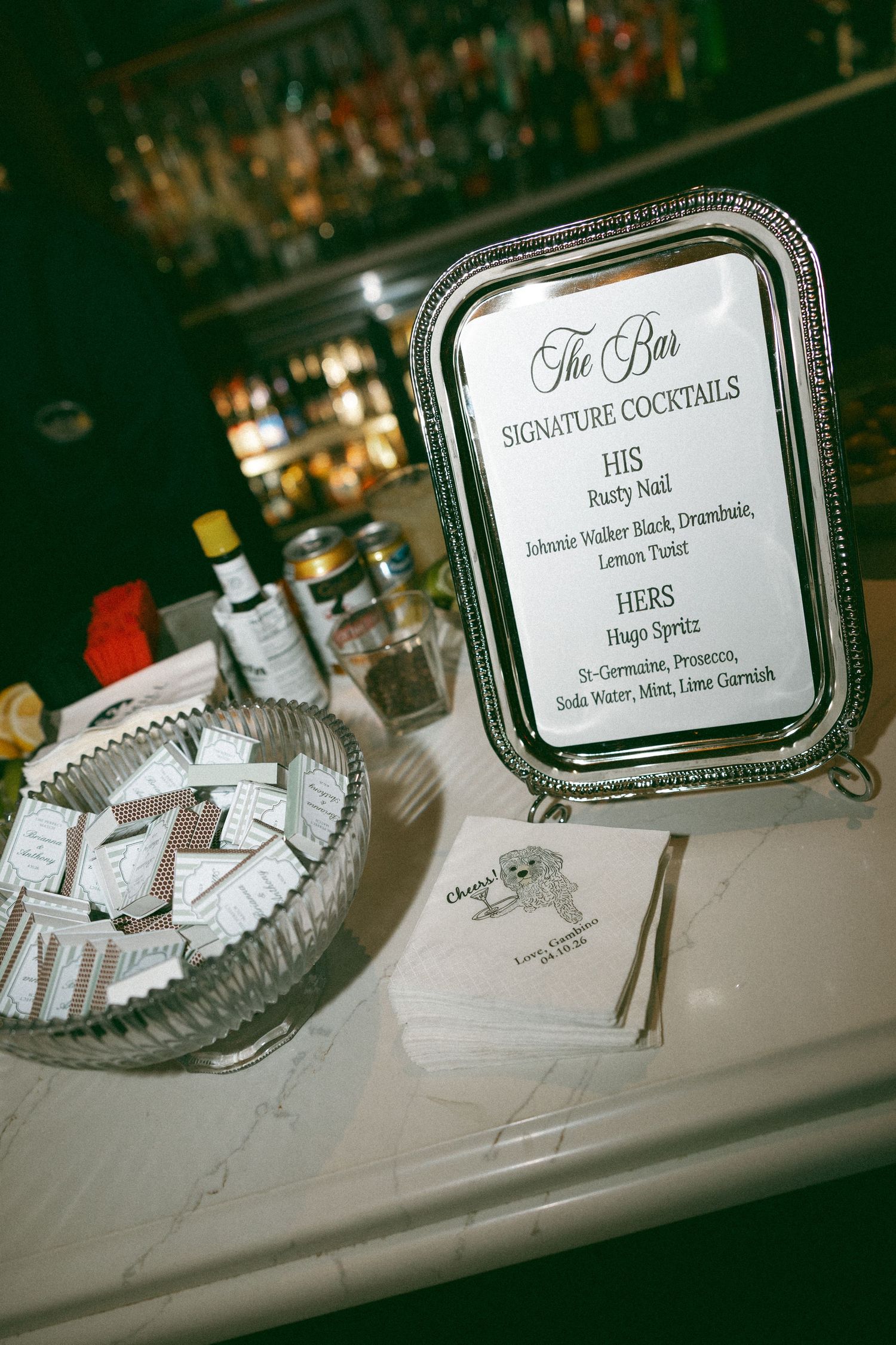 Wedding bar sign displaying signature cocktails His and Hers, displayed on silver tray with party favors nearby.