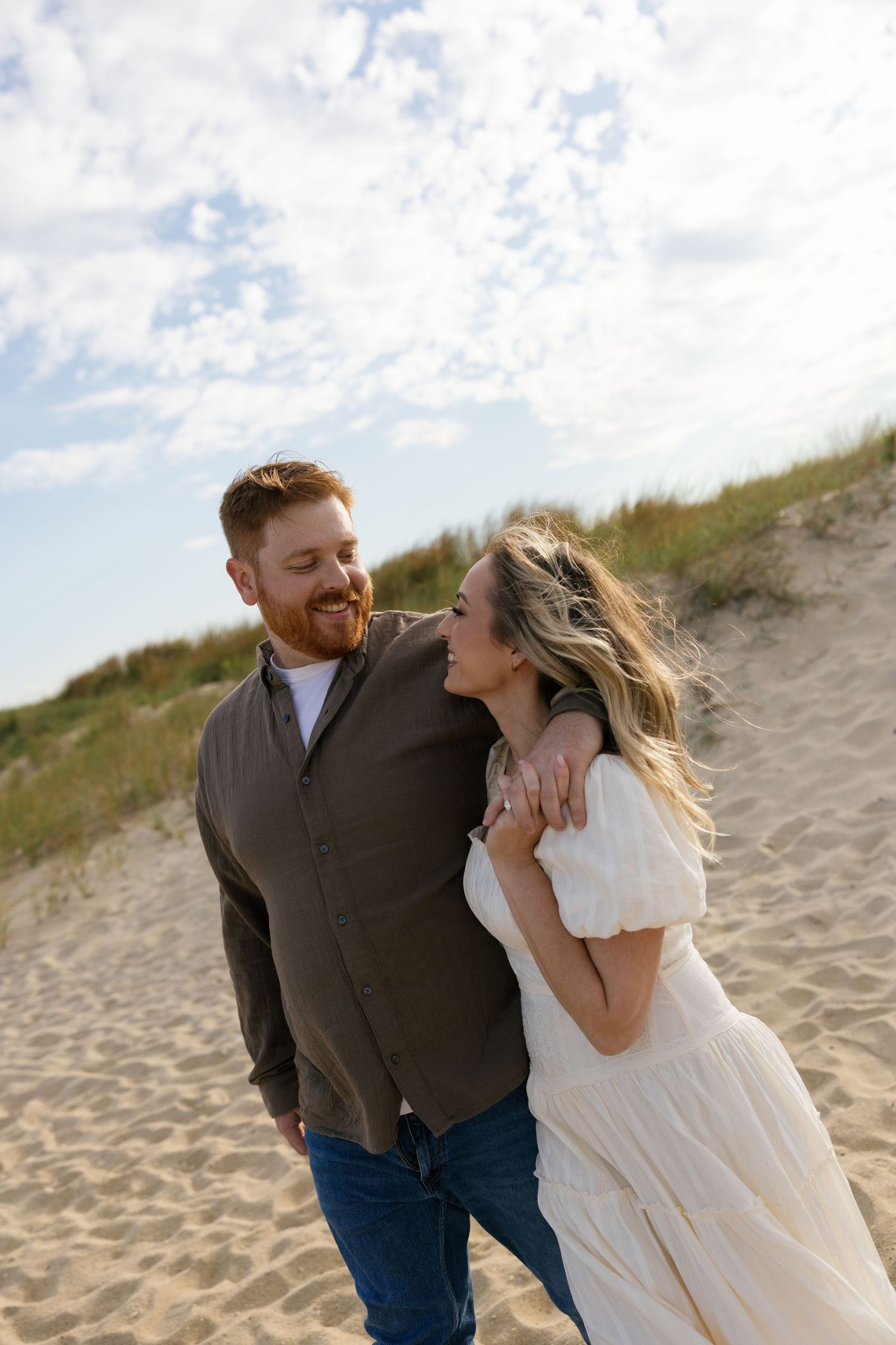 A couple shares romantic moments on a sandy beach during sunset as her white dress flows in the wind.