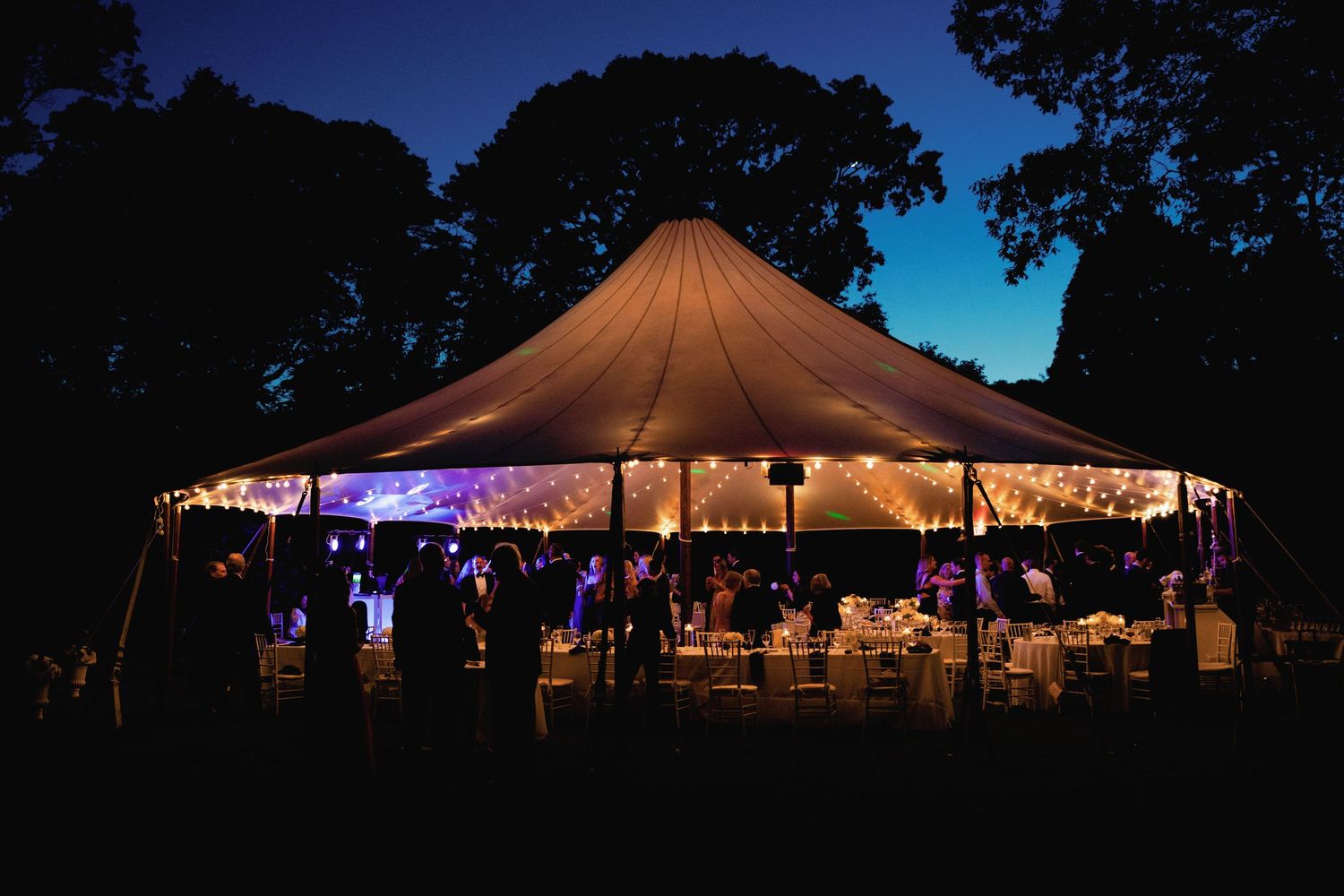 Illuminated wedding reception tent glows at dusk creating a magical outdoor atmosphere.