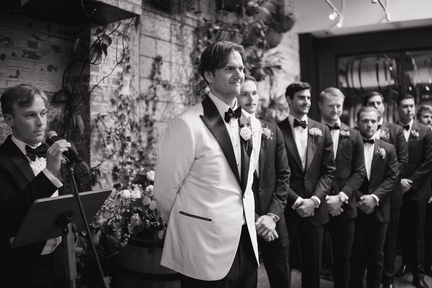 A group in formal black tuxedos poses together against a textured wall at an elegant event.
