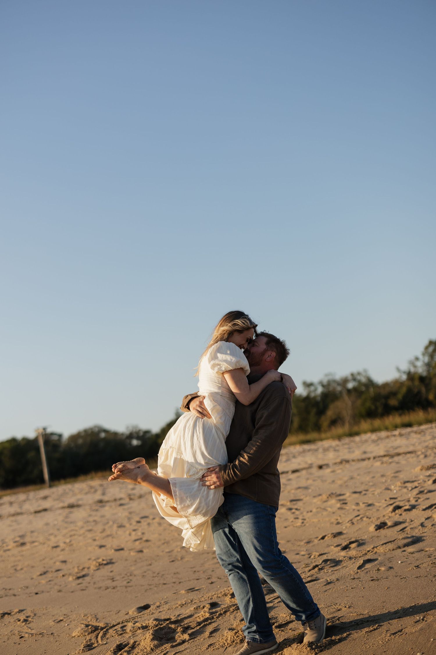 Couple in casual attire sharing romantic moments on a sandy beach under clear blue skies.