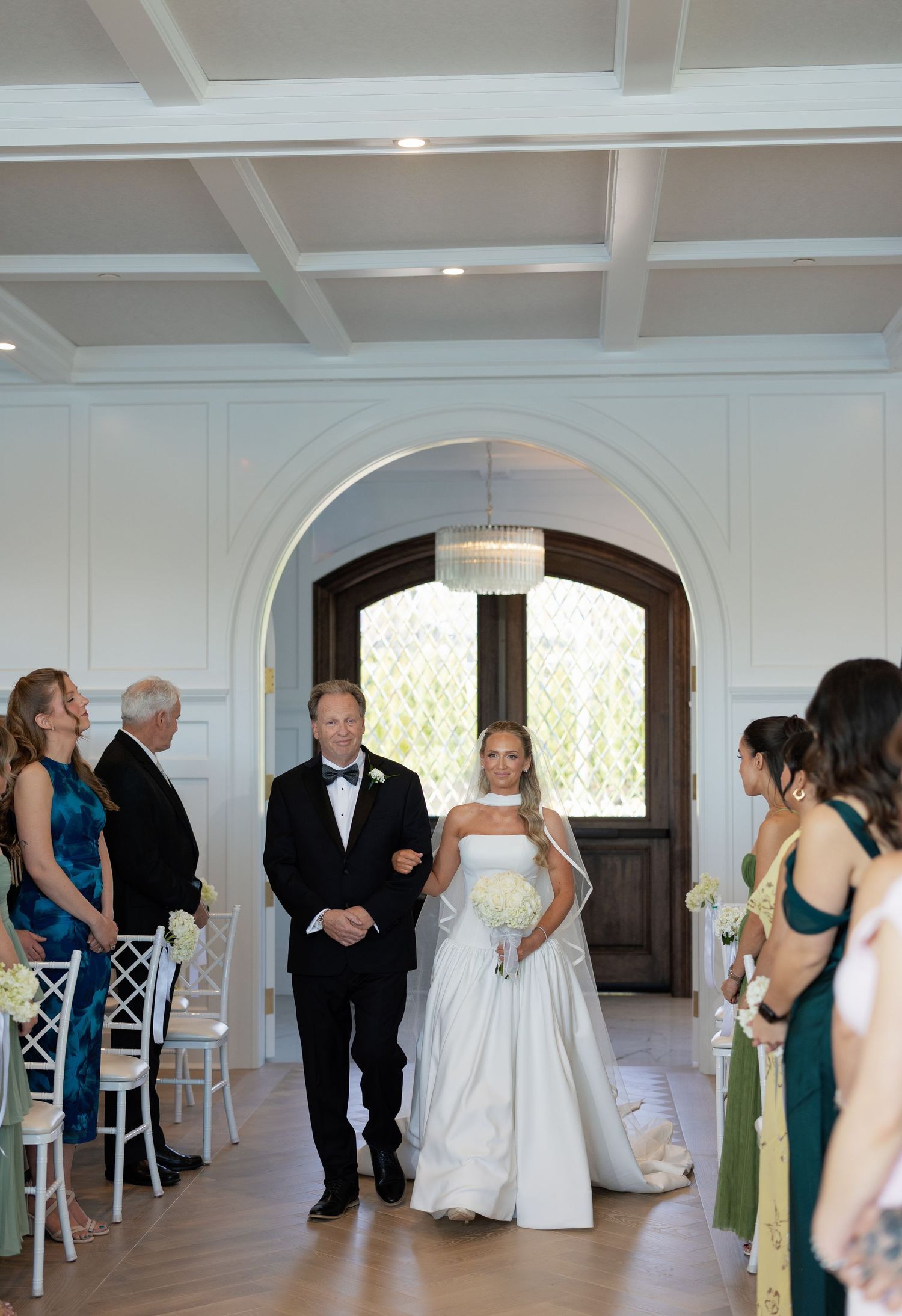Bride walking down the aisle with escort in elegant white indoor wedding ceremony venue.