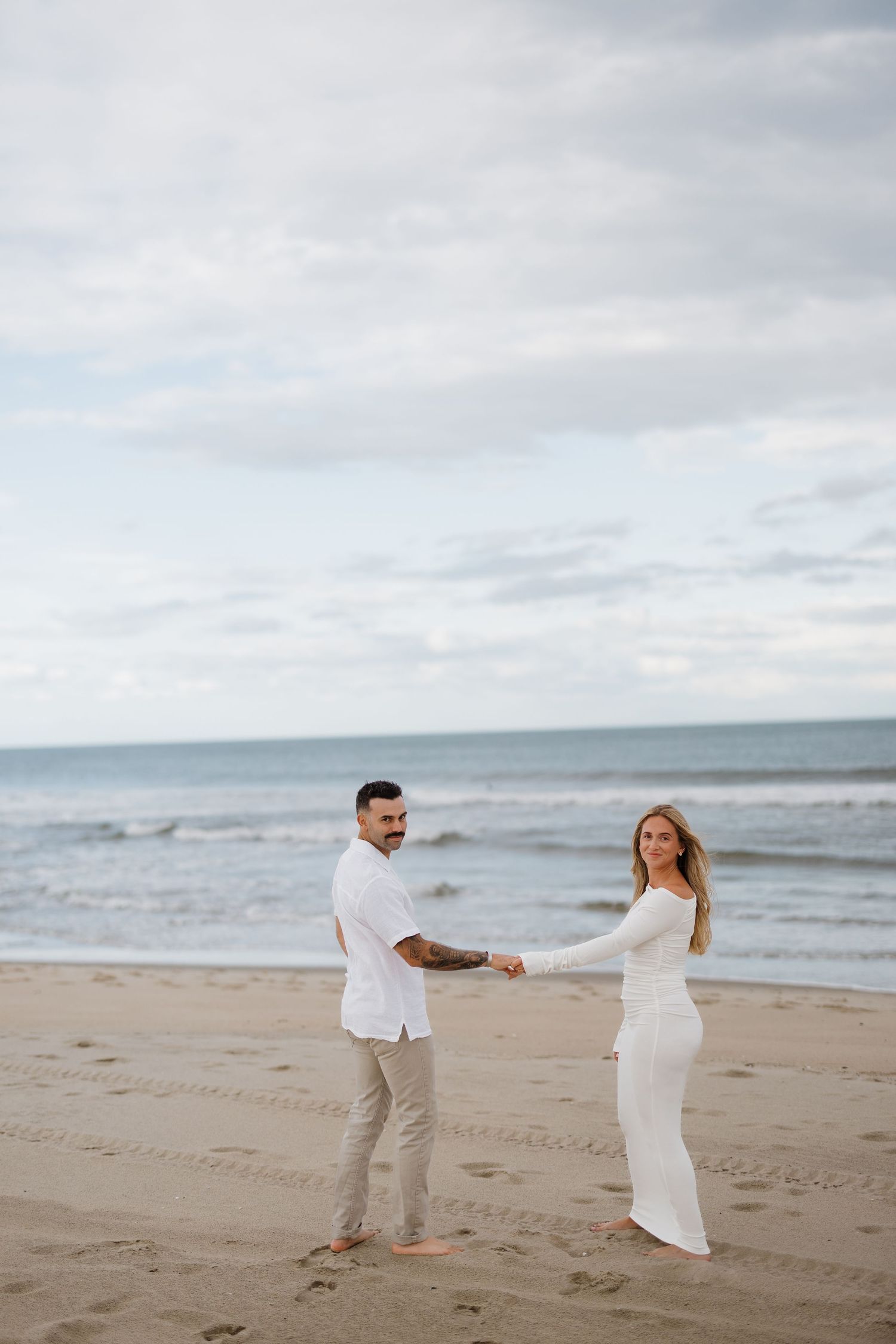 Couple in white attire walking along a sandy beach under cloudy skies.
