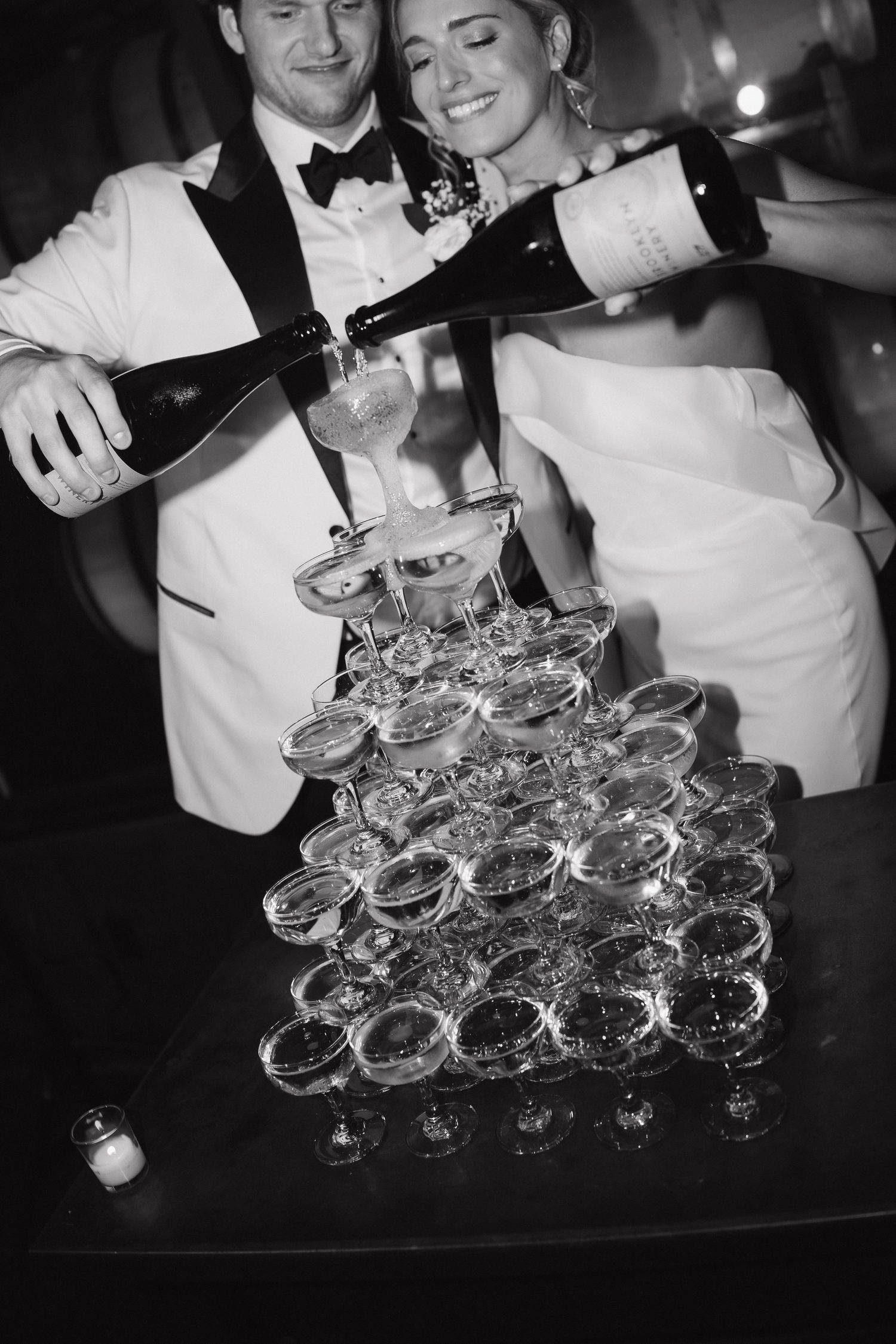 Hands pour champagne into a pyramid of crystal glasses stacked on a dark surface.