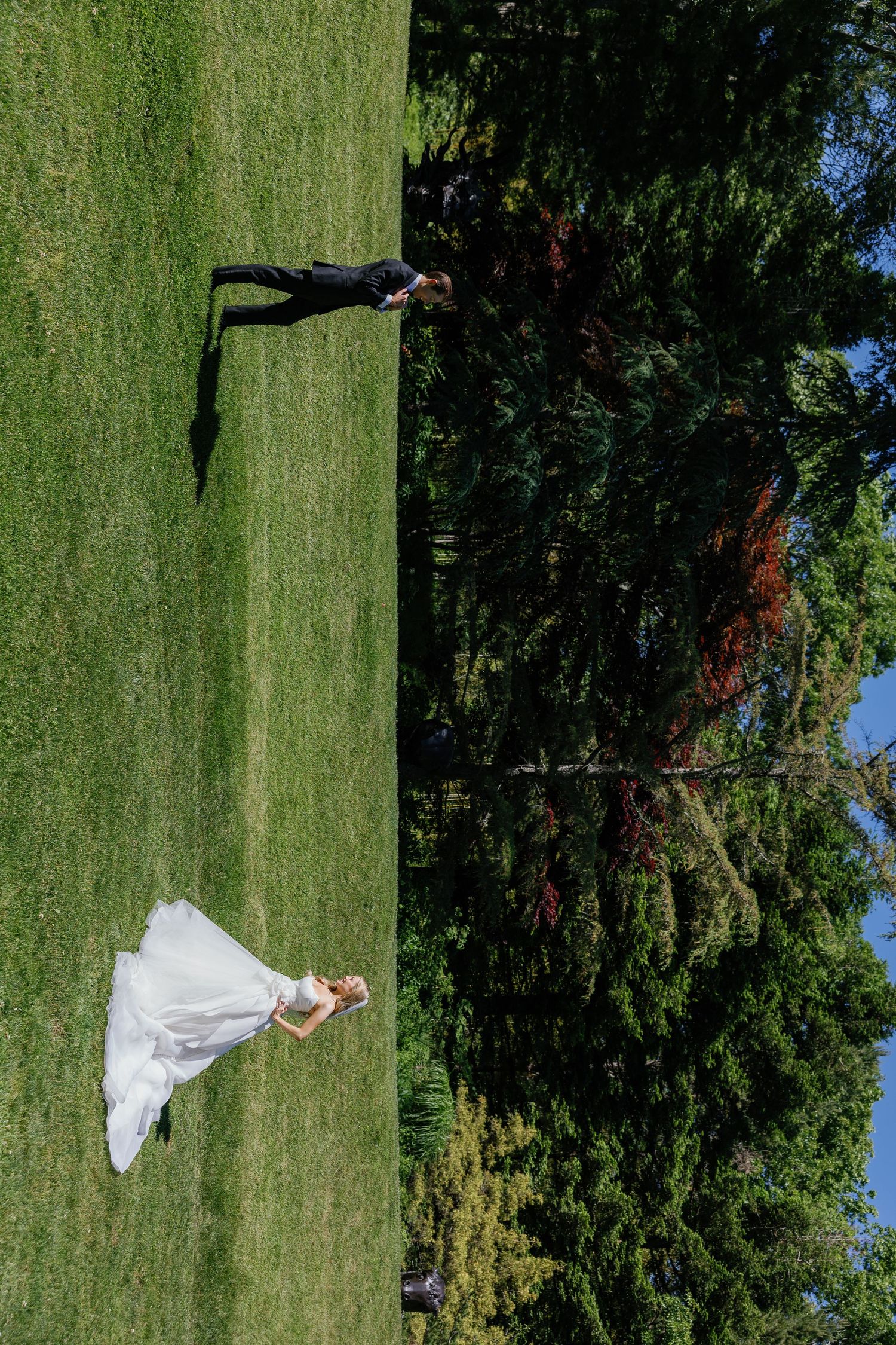 A wedding couple poses playfully on a lush green lawn with dramatic aerial views.