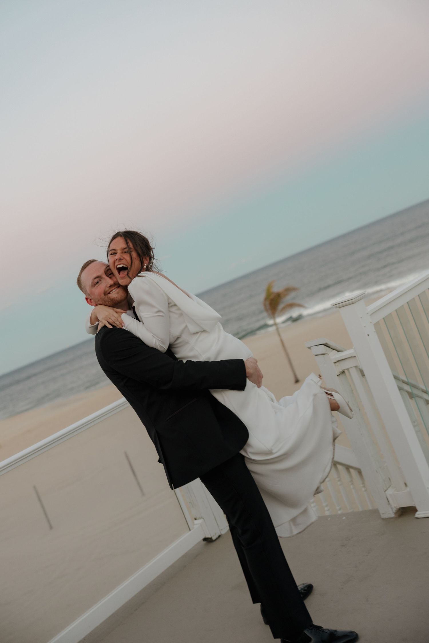Joyful bride laughing as groom lifts her on a sandy beach at sunset, both dressed in elegant wedding attire.