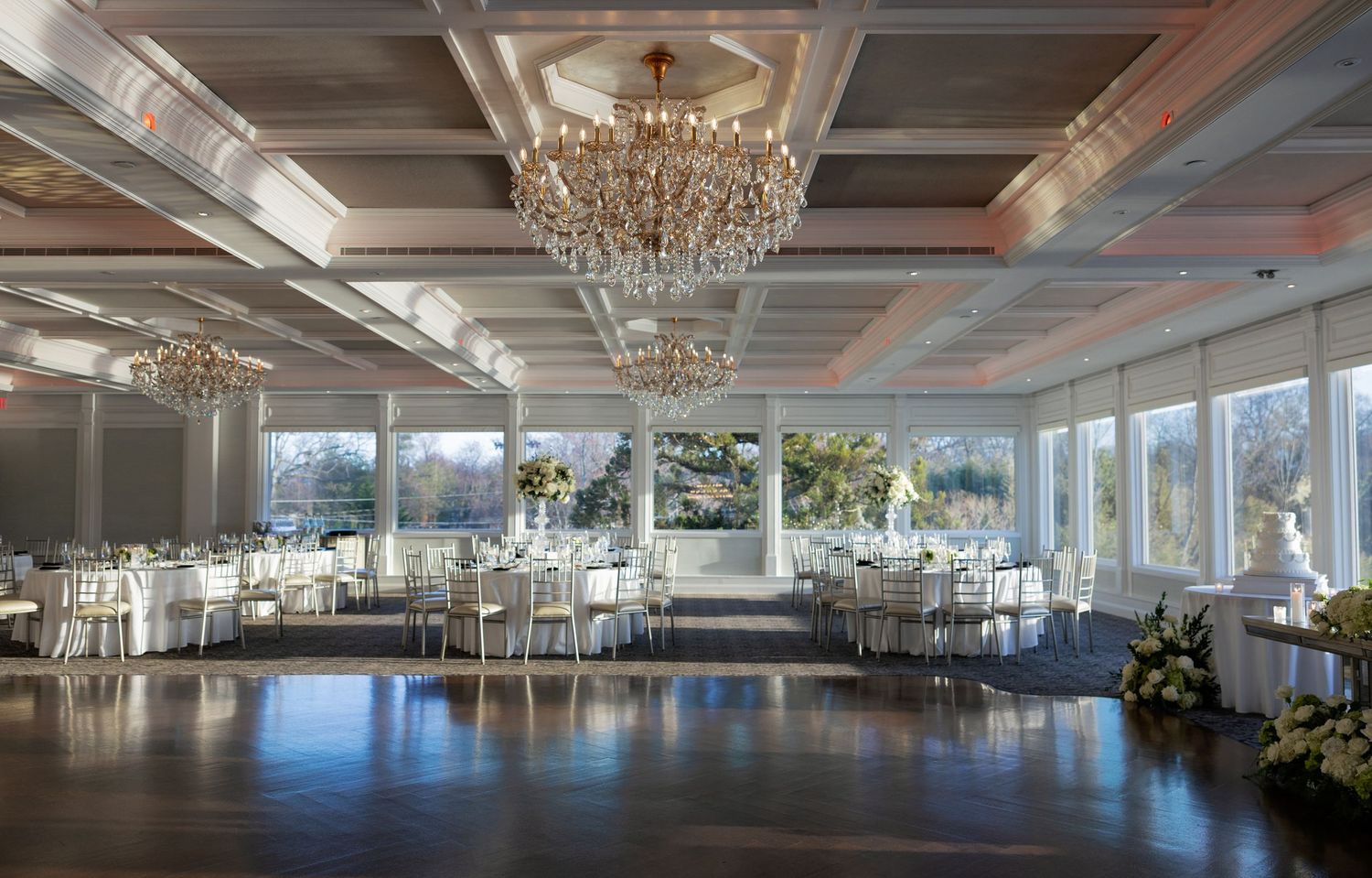 Elegant wedding reception hall with crystal chandeliers, white round tables, gold chairs, and floor-to-ceiling windows.