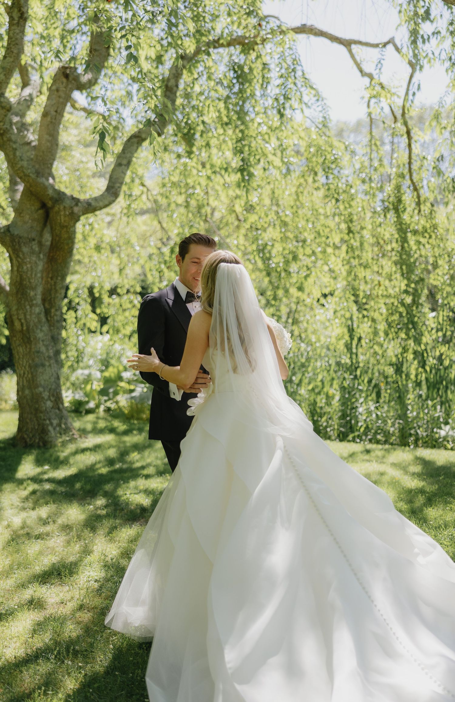 A romantic wedding couple embraces under weeping willow trees in a sunlit garden setting.