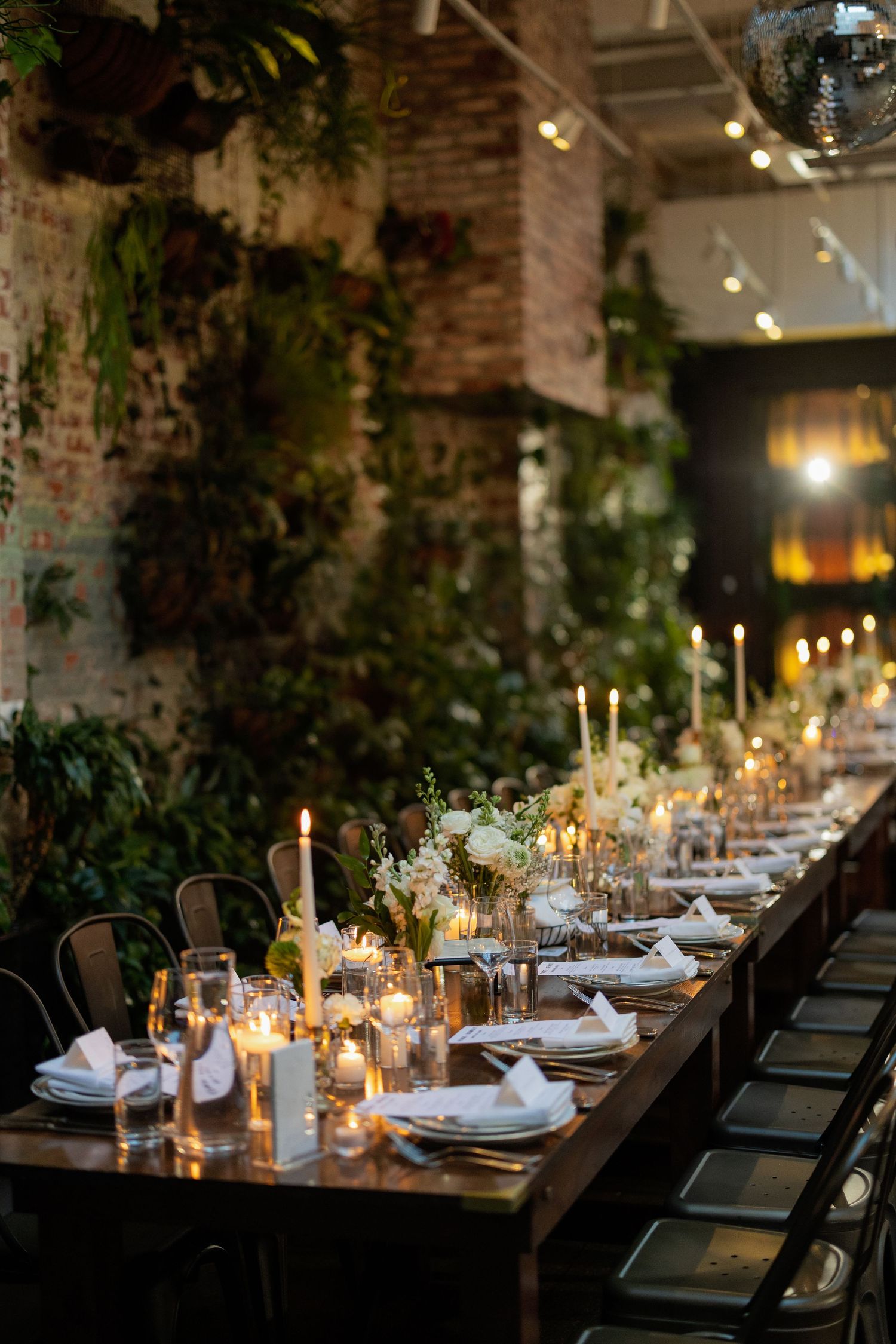 Elegant wedding reception table with candlelight, white floral centerpieces and exposed brick walls adorned with greenery.