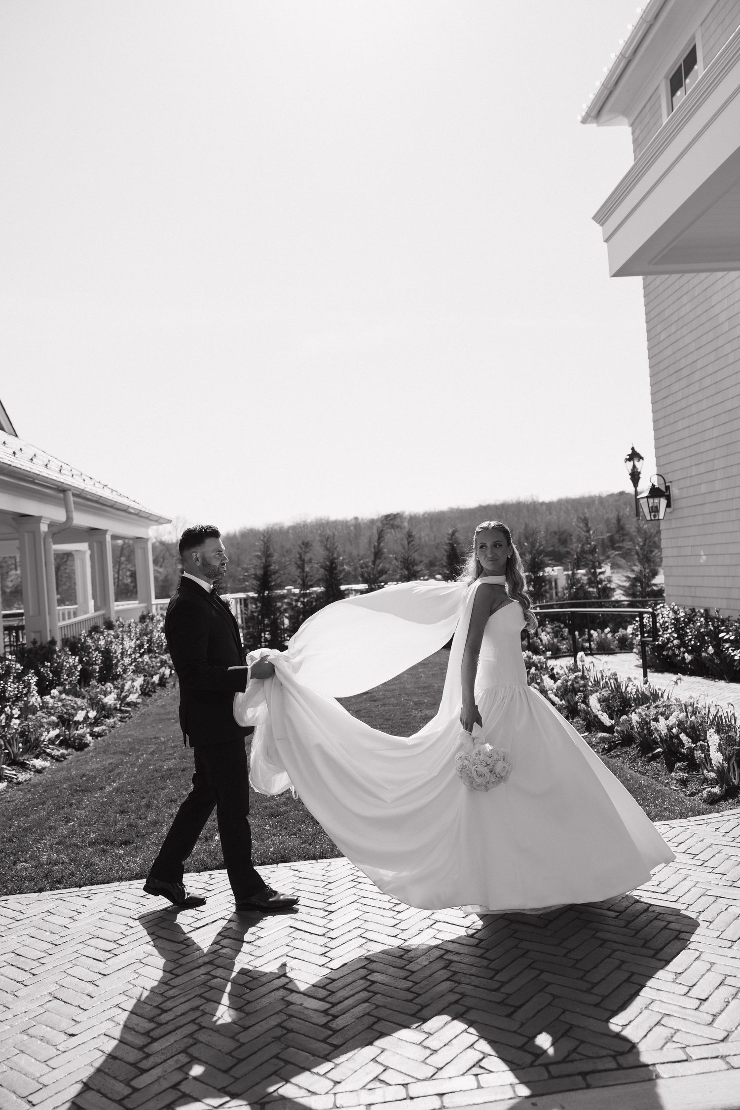 Black and white photo of bride and groom walking outdoors, wedding dress flowing in the breeze.