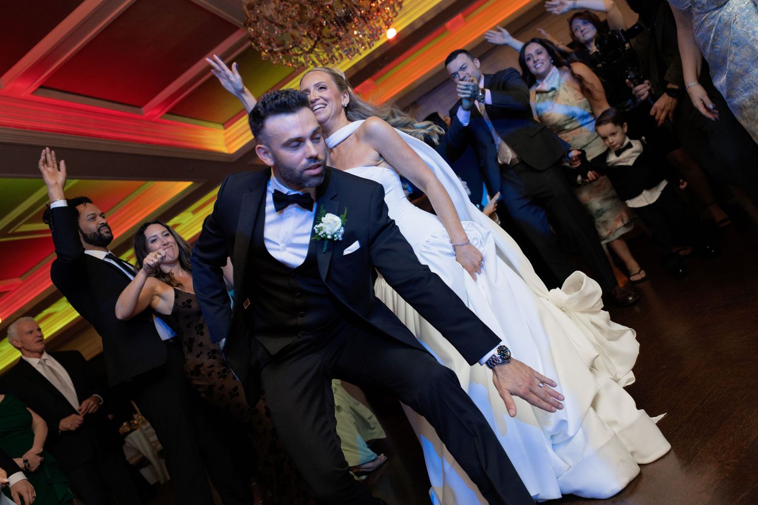 Energetic bride and groom dancing joyfully with wedding guests on colorfully lit reception dance floor.