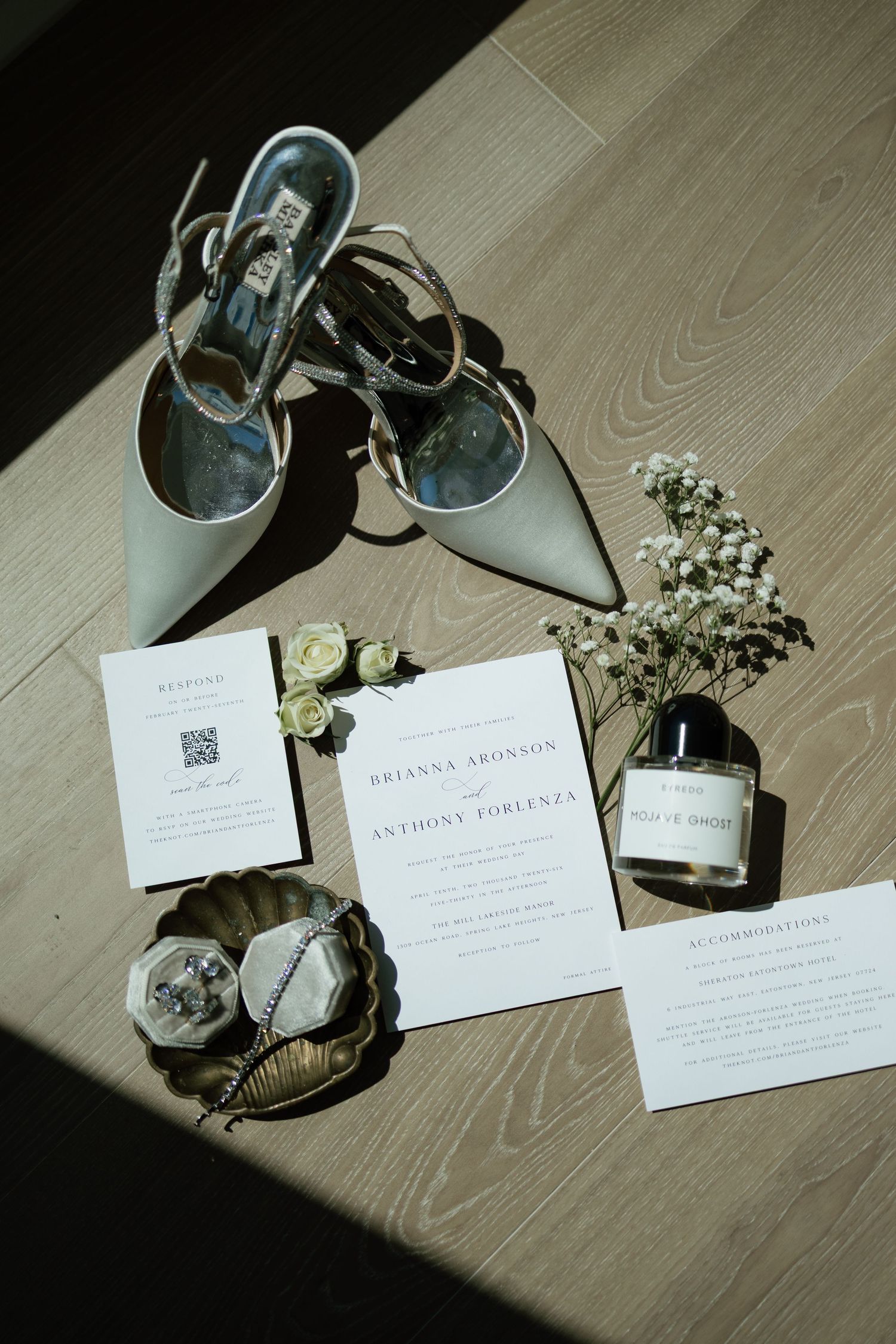 Elegant wedding flat lay featuring bridal heels, invitations, perfume, and floral details on wood floor.