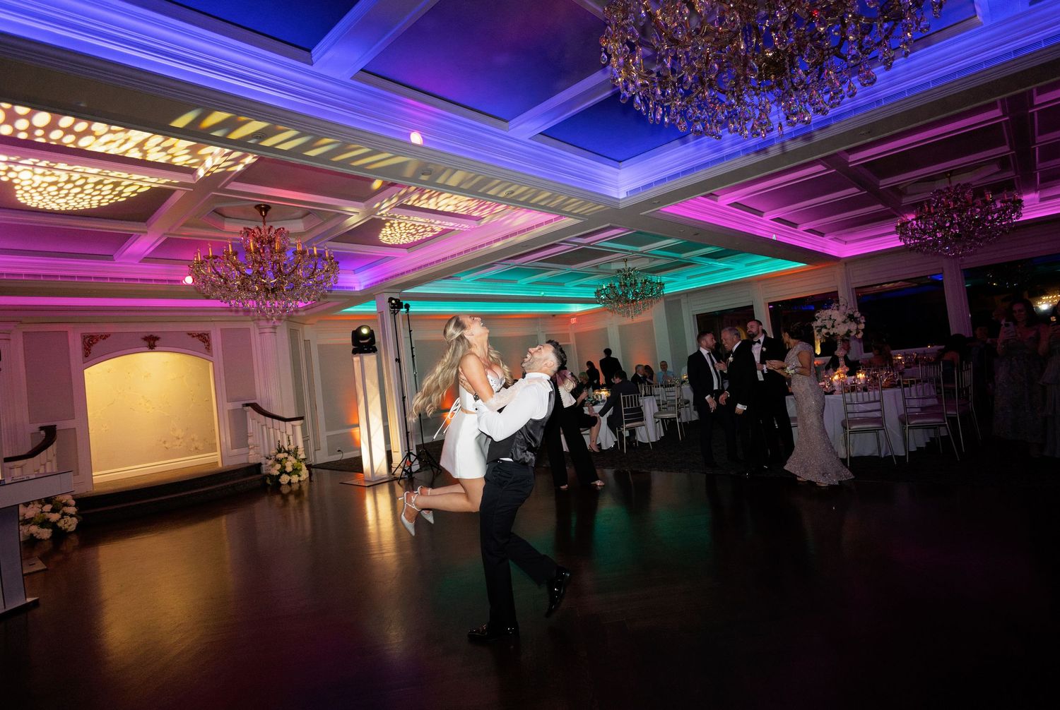 Bride and groom share first dance in ornate ballroom with colorful purple and teal uplighting.