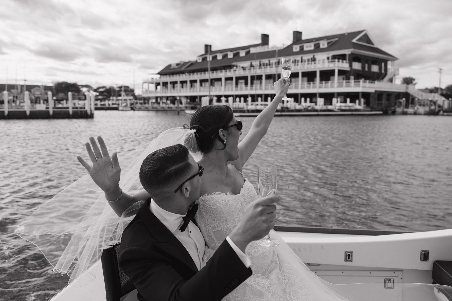Black and white photo of a bride and groom celebrating on a boat with champagne, waterfront venue in background.