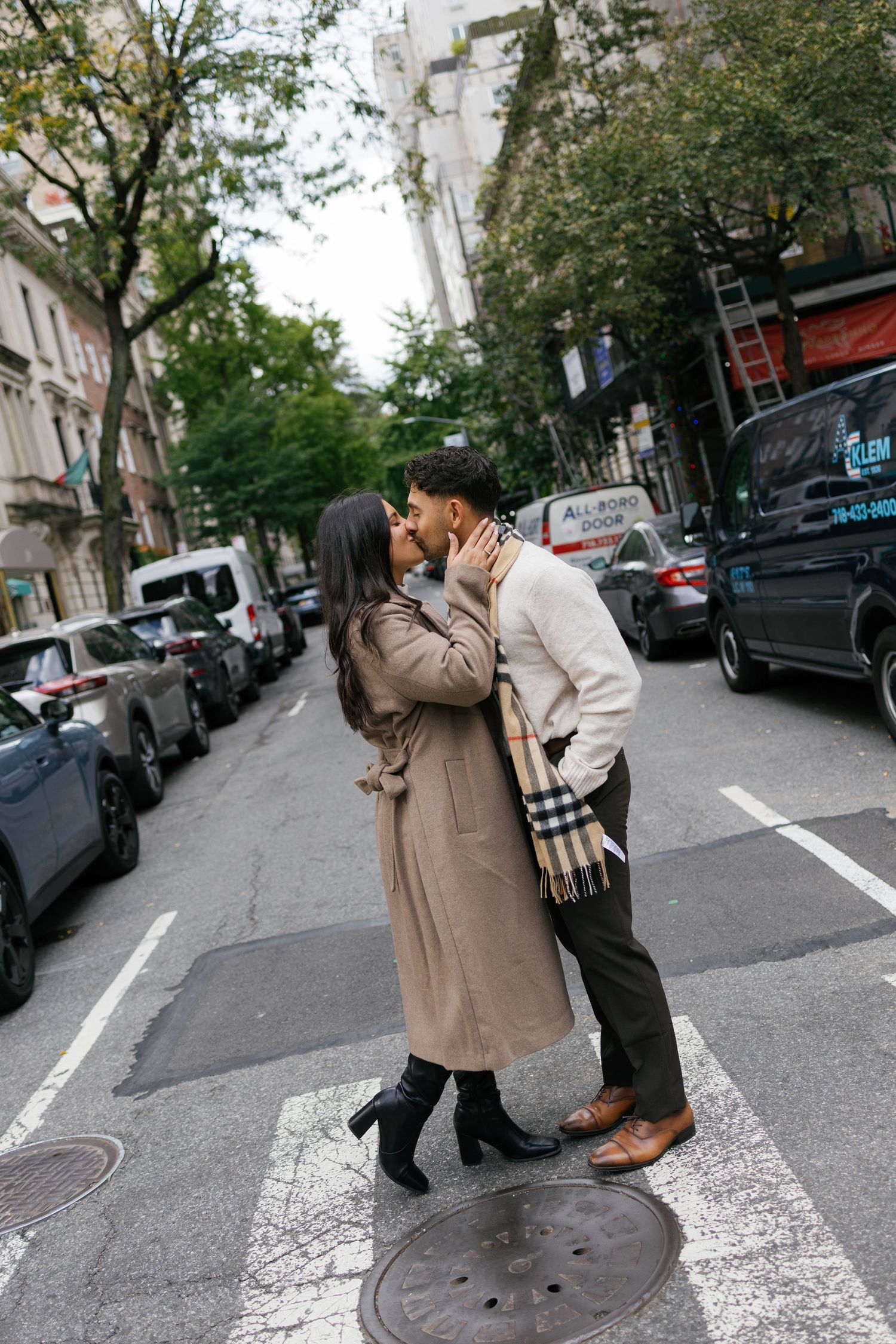A couple embraces on a quiet city street with parked cars and buildings in the background.