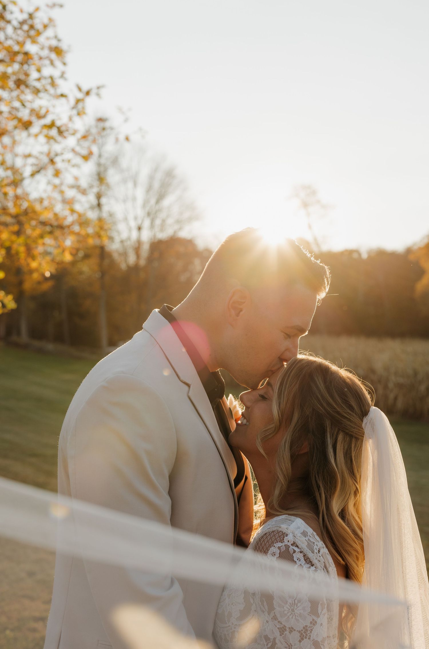 Romantic couple embraces in golden sunset light with autumn trees and a flowing wedding veil in soft focus.