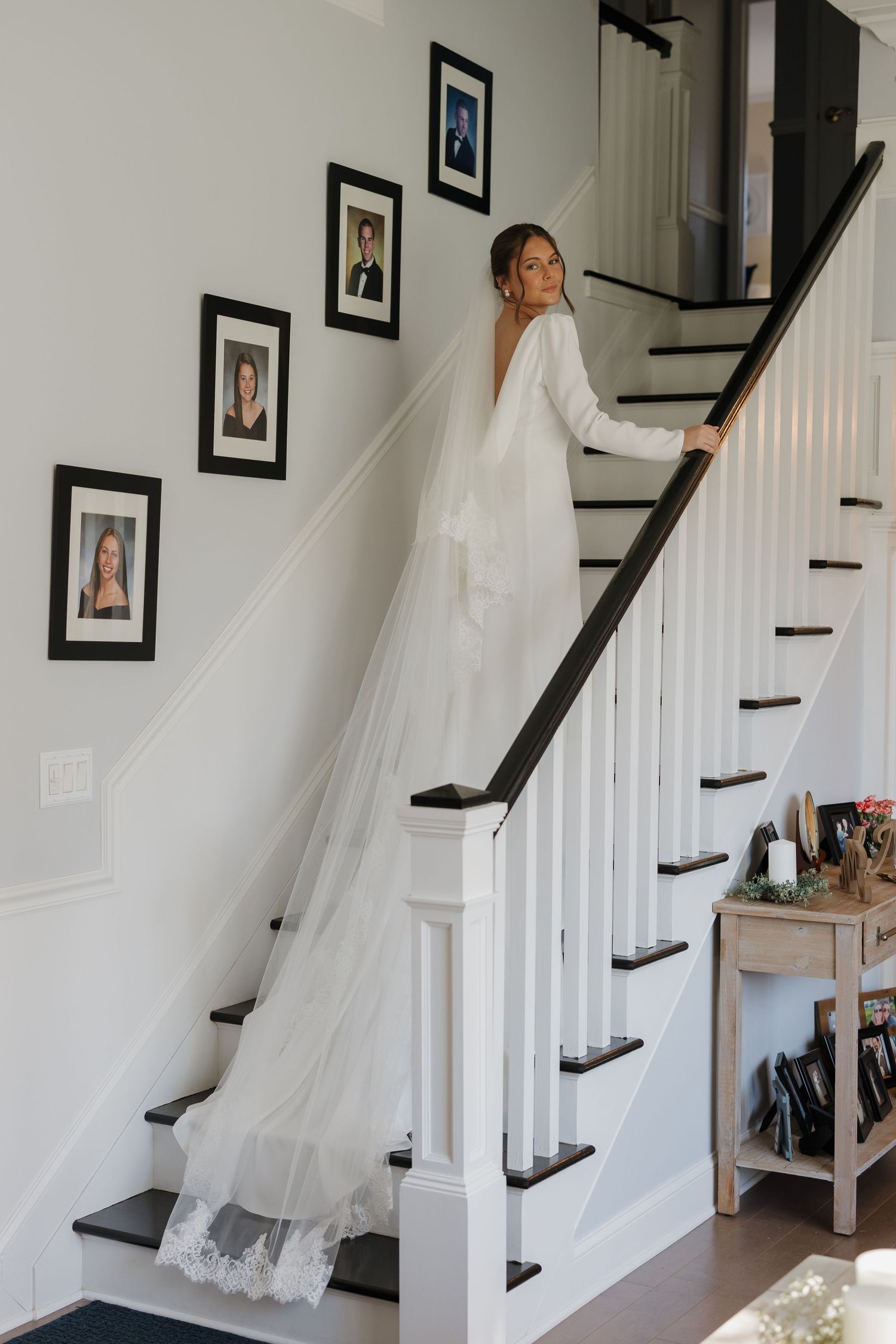 A bride in a flowing white wedding gown and long veil poses on a white staircase with framed photos on the wall.