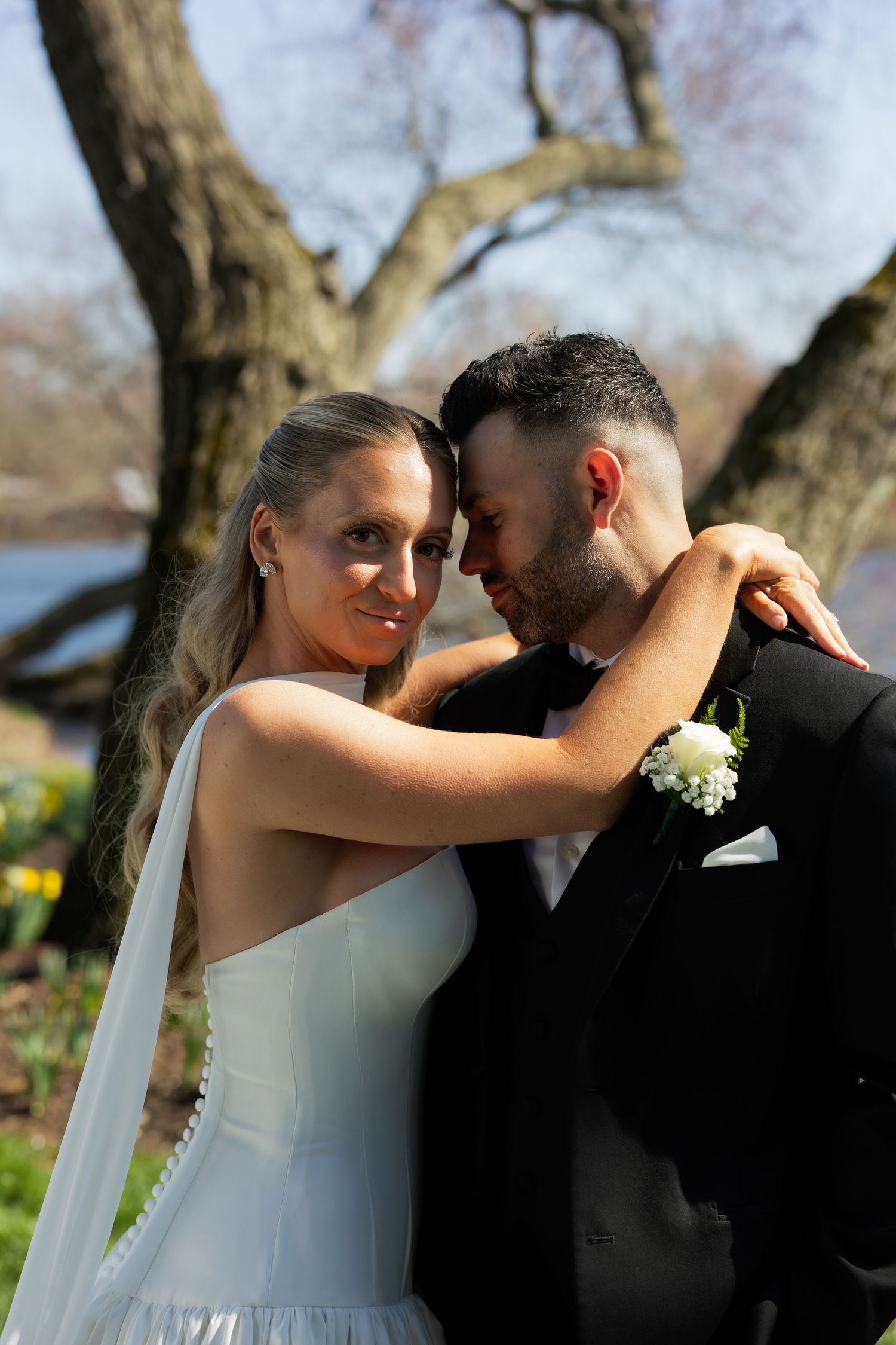 Bride and groom embrace outdoors near a tree, bride in white gown with veil smiling at camera.