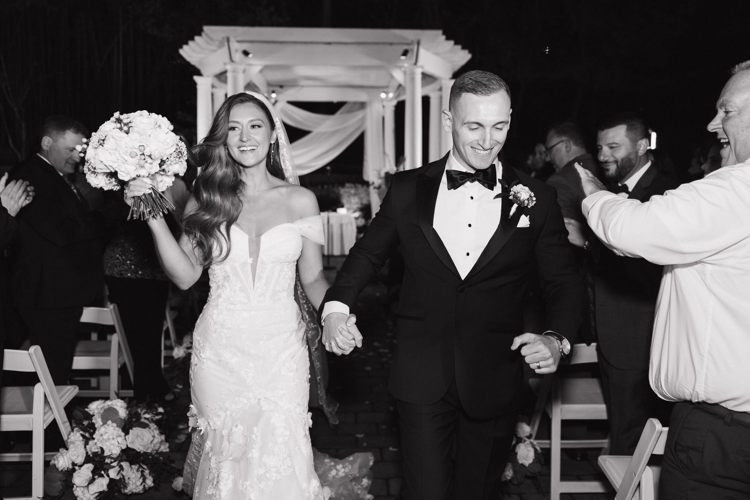 A joyful newlywed couple walks down the aisle after their ceremony while guests celebrate their union in black and white.