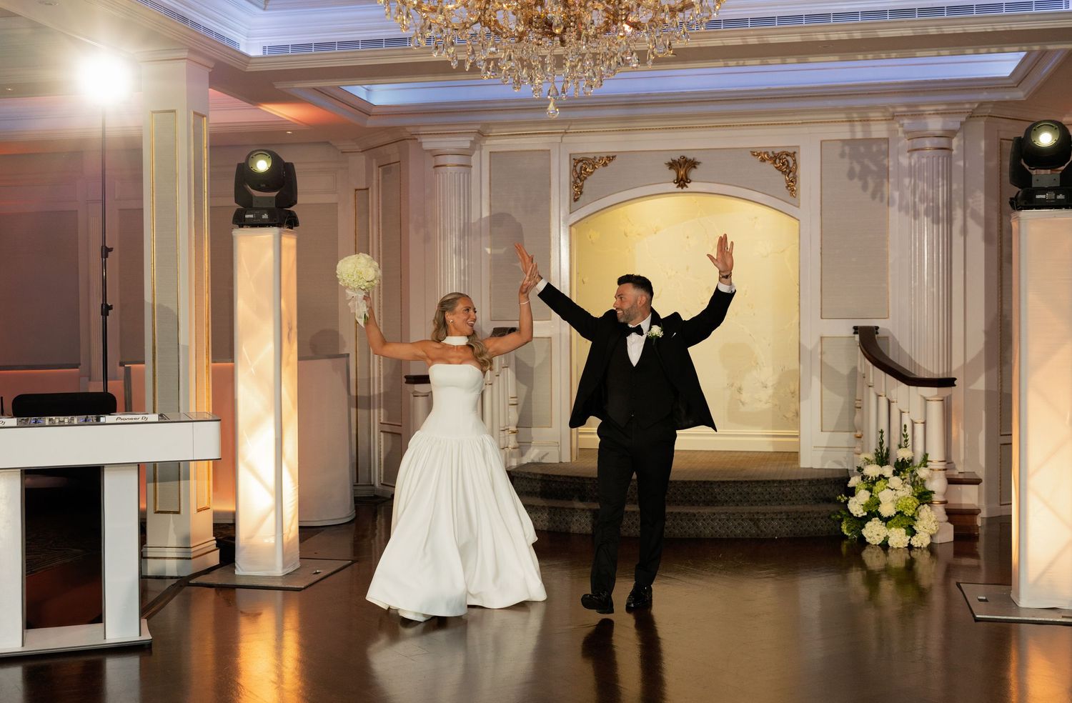 Bride and groom make grand entrance into elegant ballroom reception venue with chandelier.