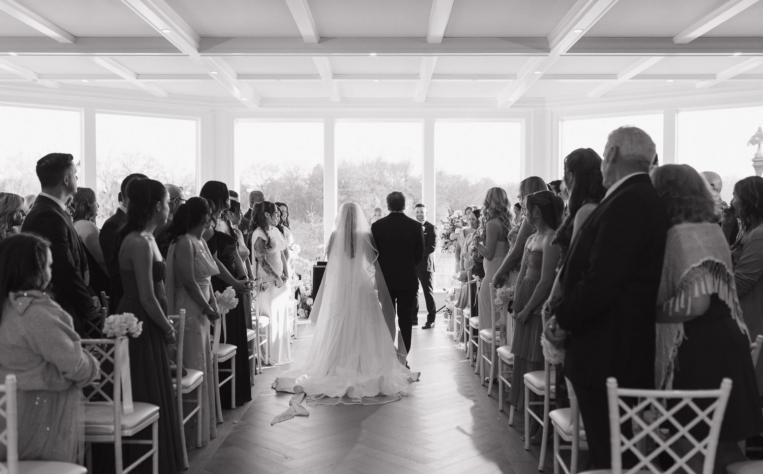Black and white photo of a bride walking down the aisle with guests seated on both sides at an elegant indoor wedding.