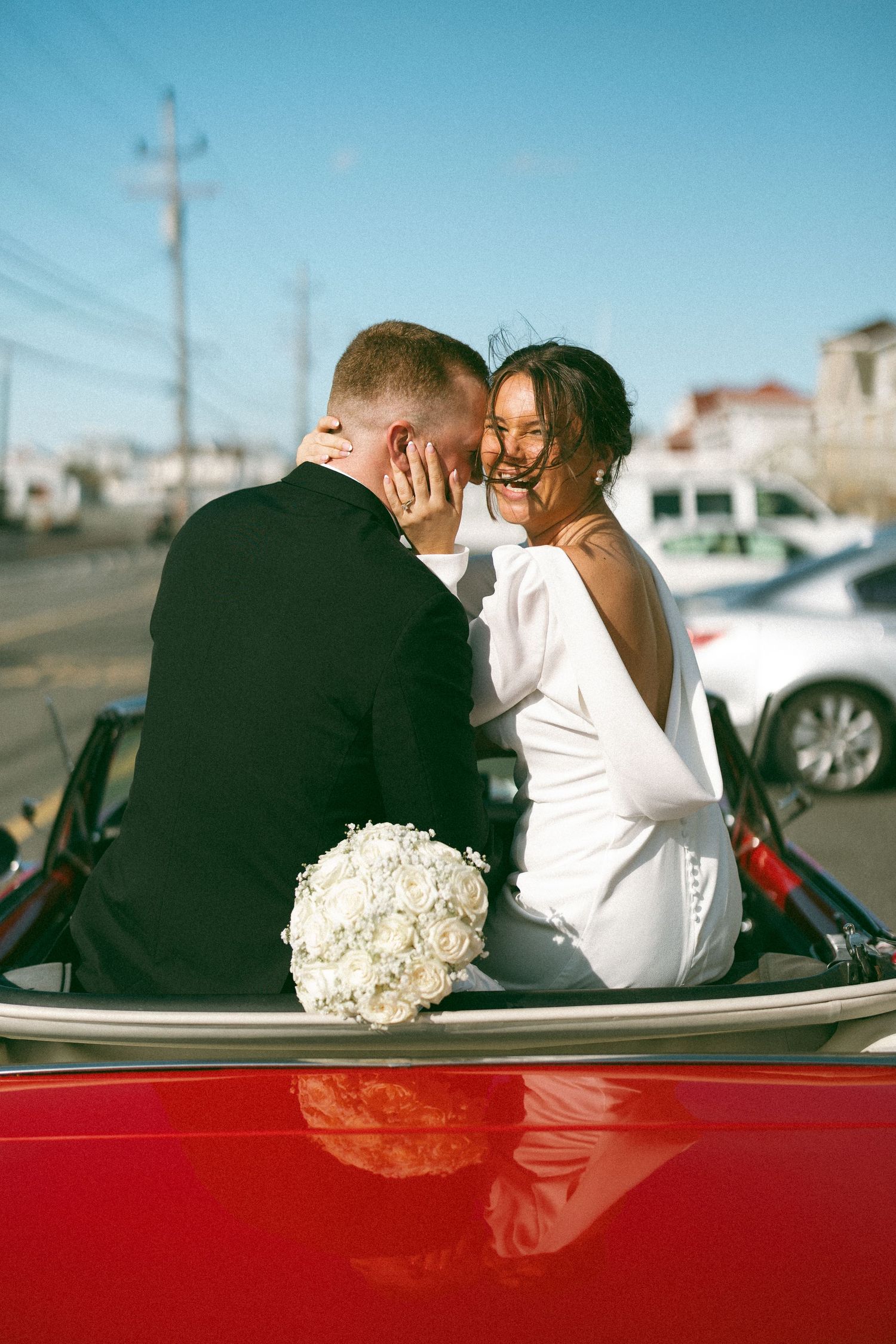 A couple shares an intimate moment in a red convertible car with a white bouquet of flowers on their wedding day.