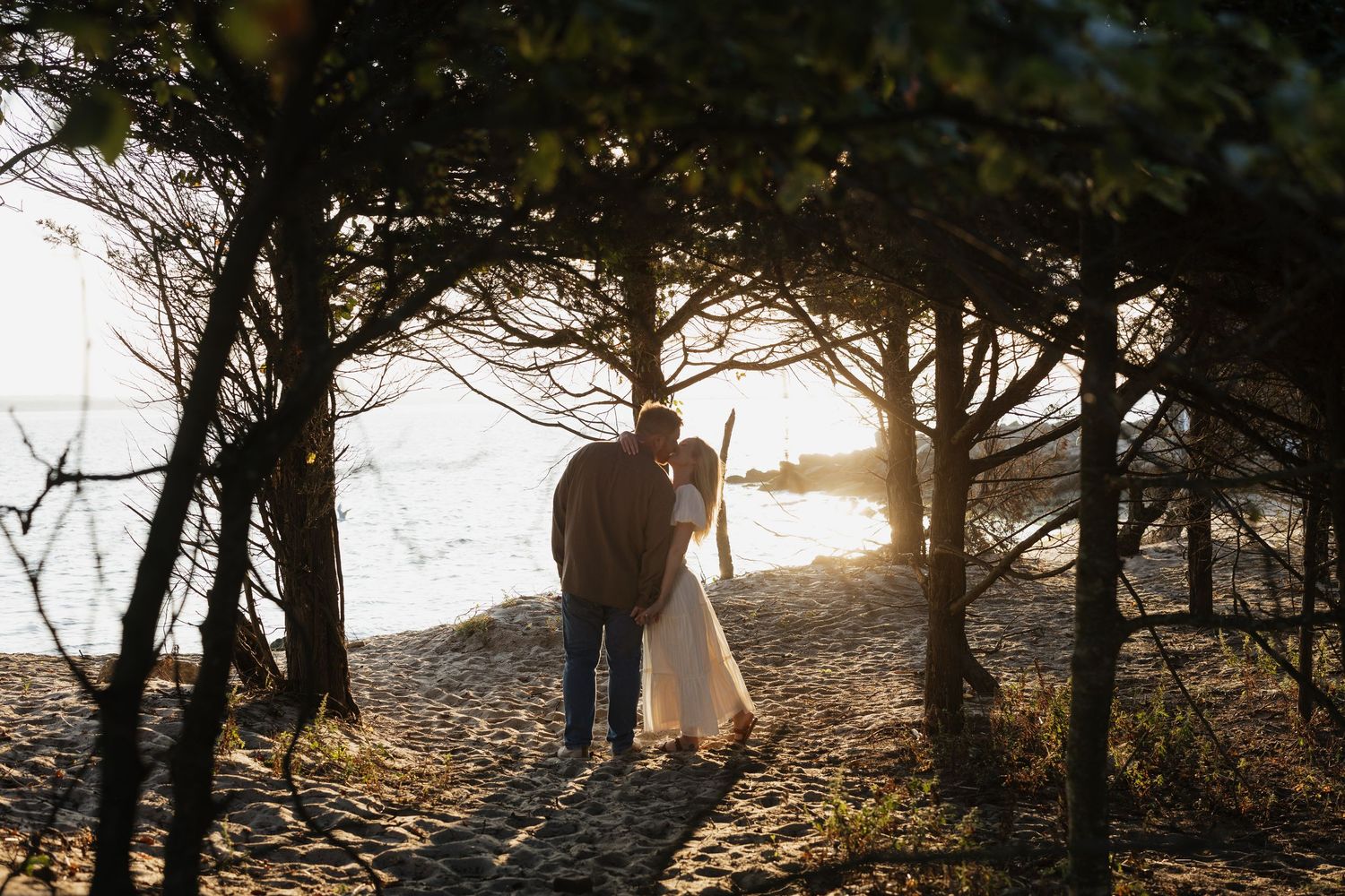 A couple stands silhouetted against a golden sunset on a rocky beach path framed by trees.
