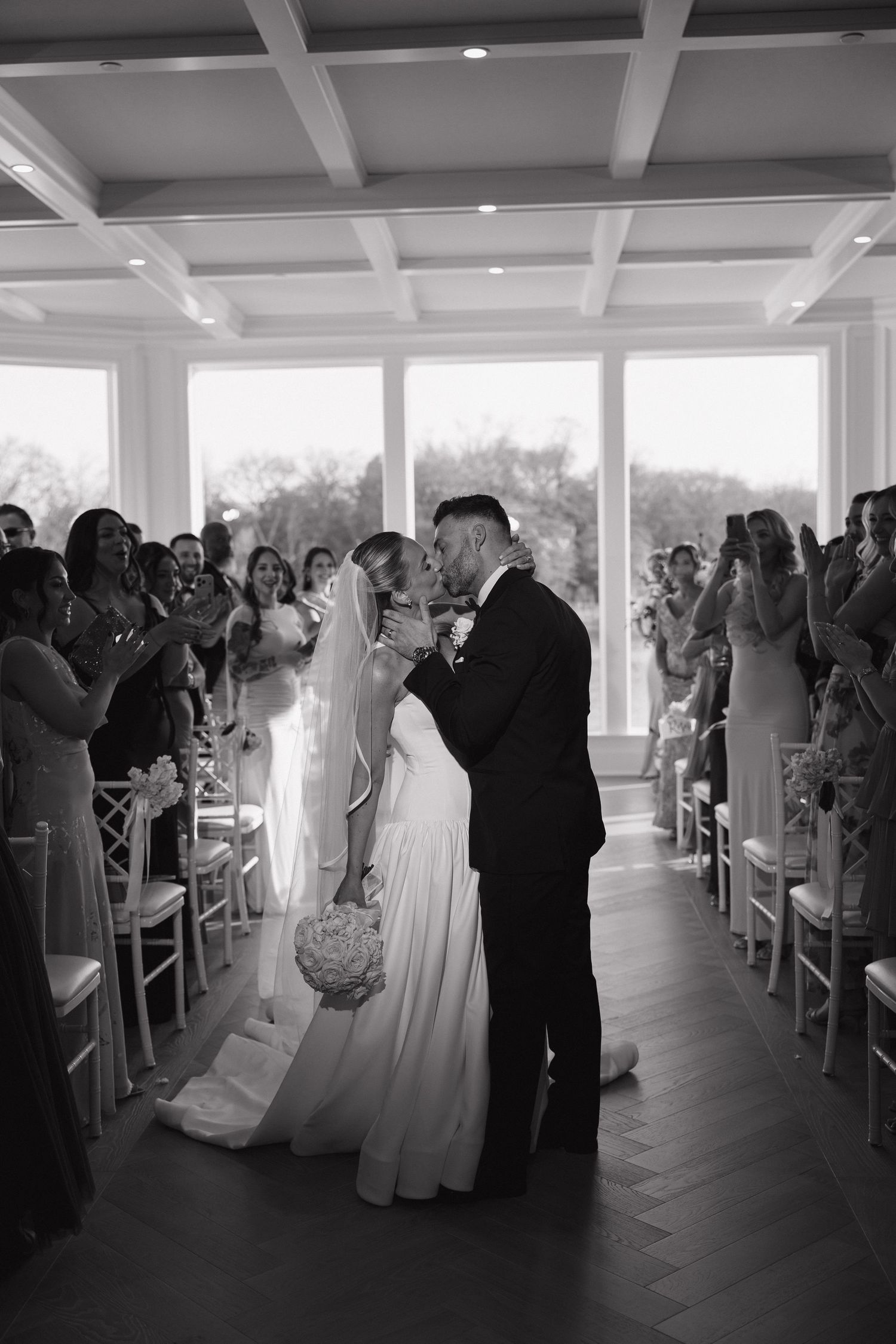 Black and white photo of a bride and groom sharing their first kiss at the altar during an indoor wedding ceremony.
