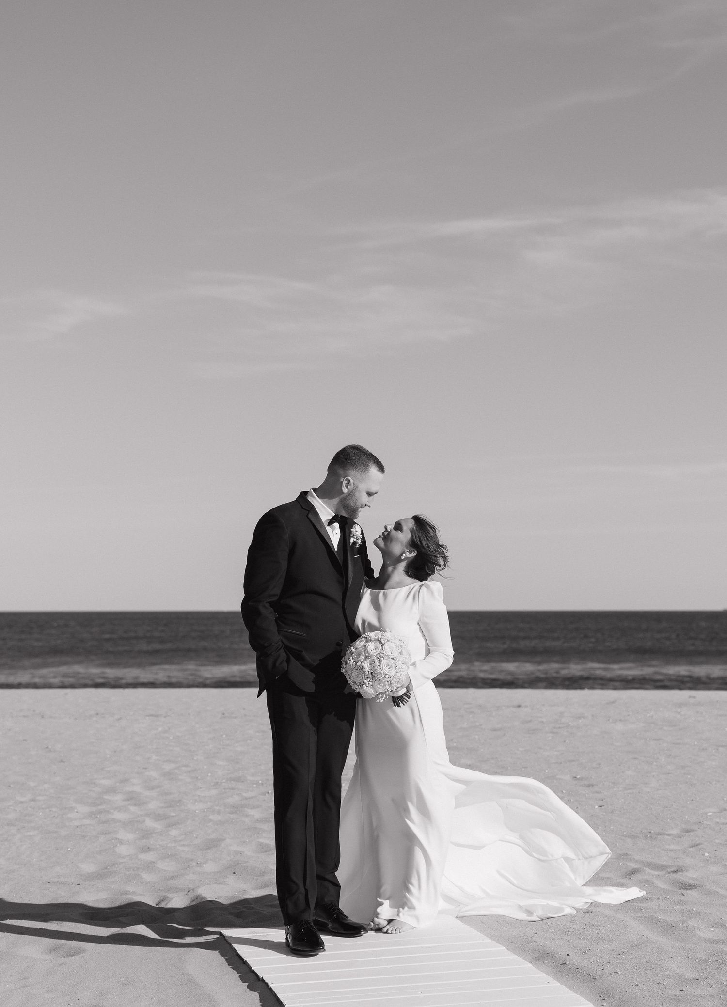 A romantic black and white beach wedding photo of a couple embracing on a boardwalk by the ocean.