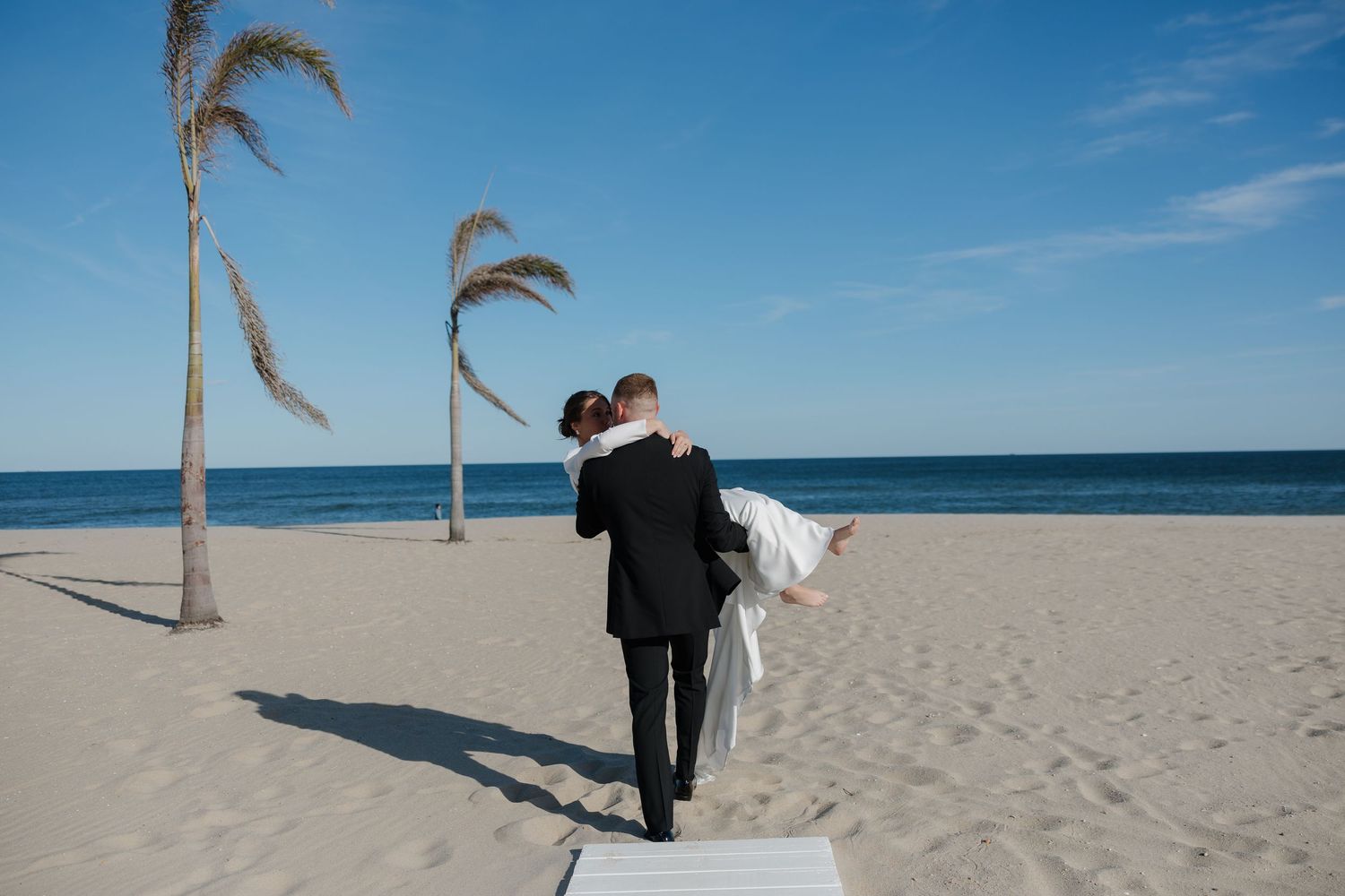 A couple in formal wedding attire embraces on a sunny beach with palm trees and blue ocean in the background.