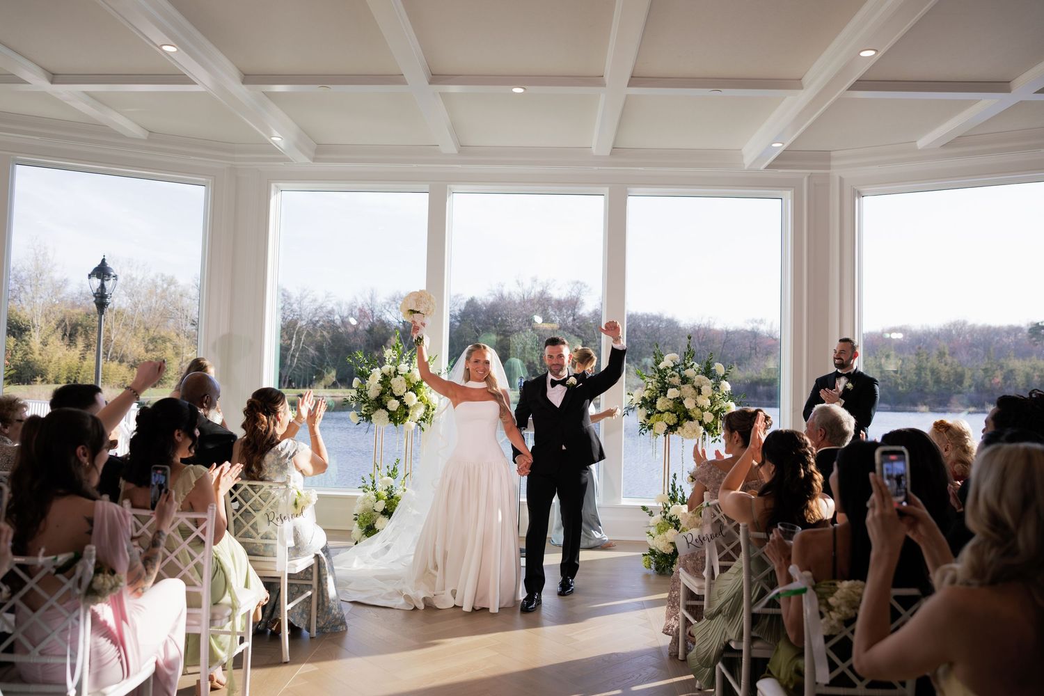Bride and groom celebrate at wedding ceremony in elegant waterfront venue with large windows and white floral decor.