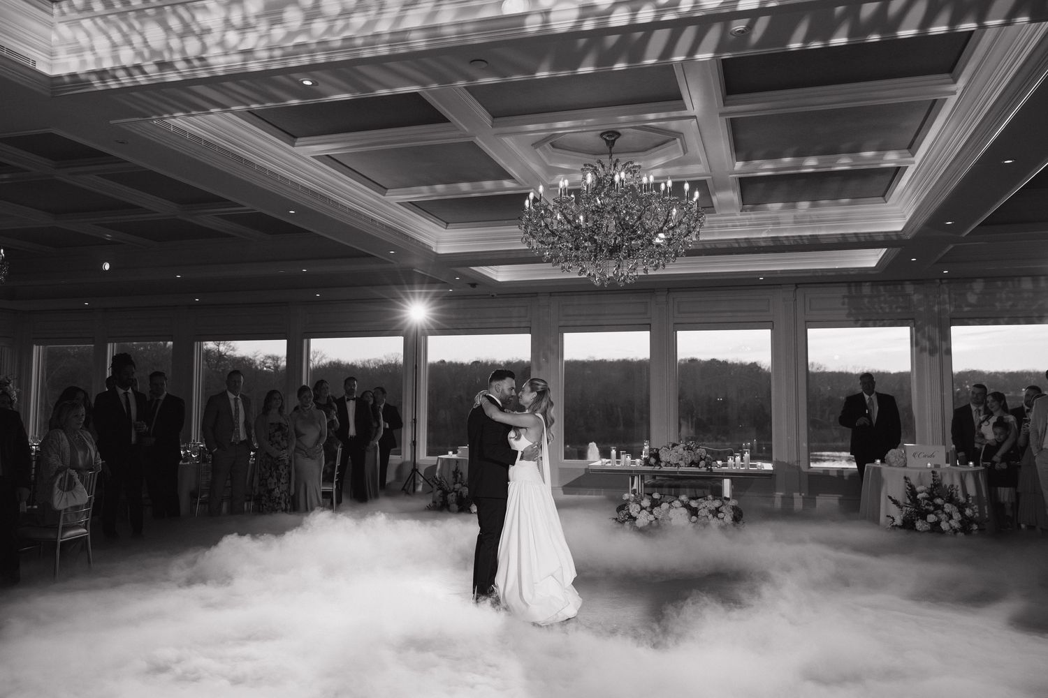 Bride and groom share first dance surrounded by fog on elegant ballroom floor with chandelier and guests watching.