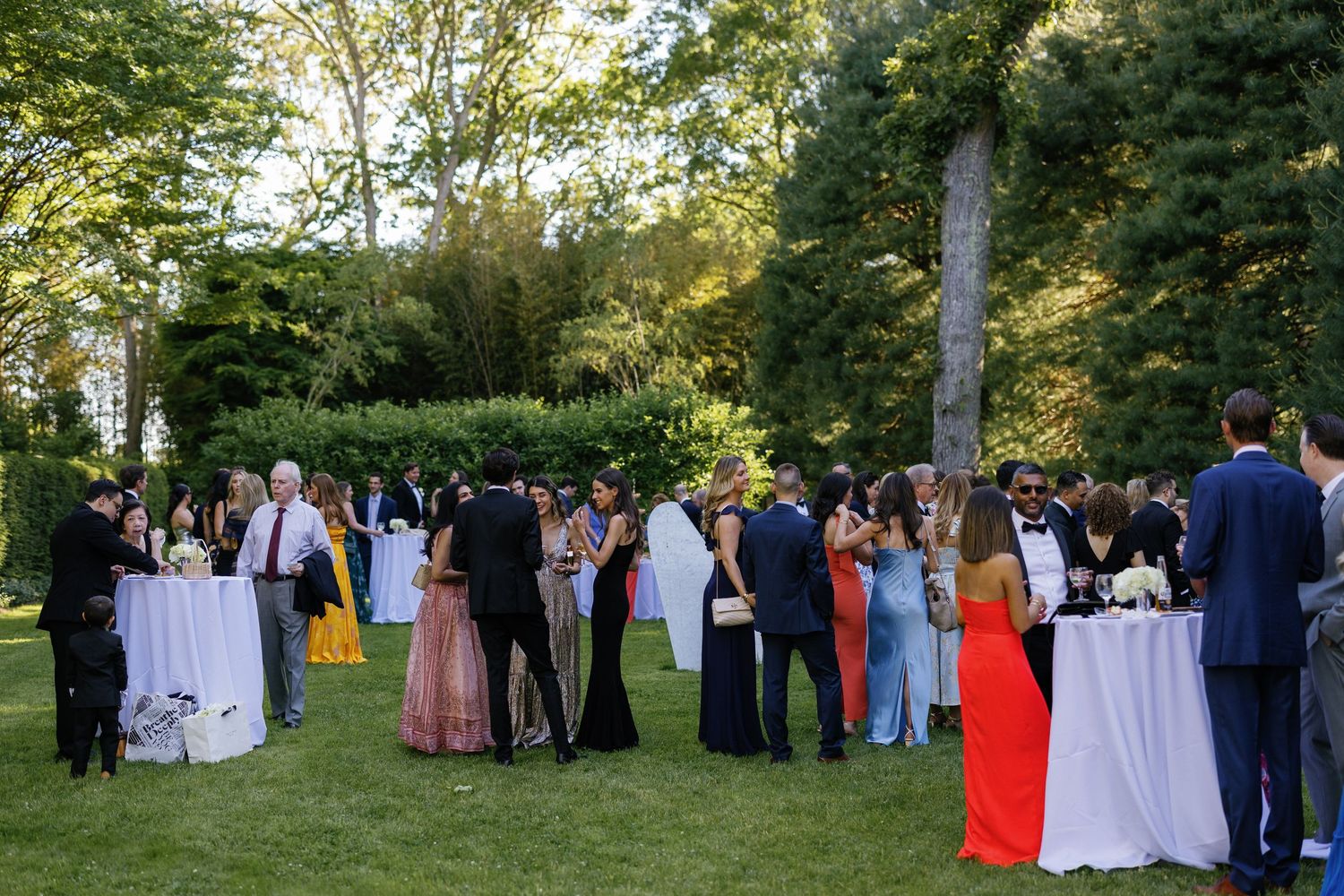 Wedding guests gather on a green lawn surrounded by trees and natural landscaping.
