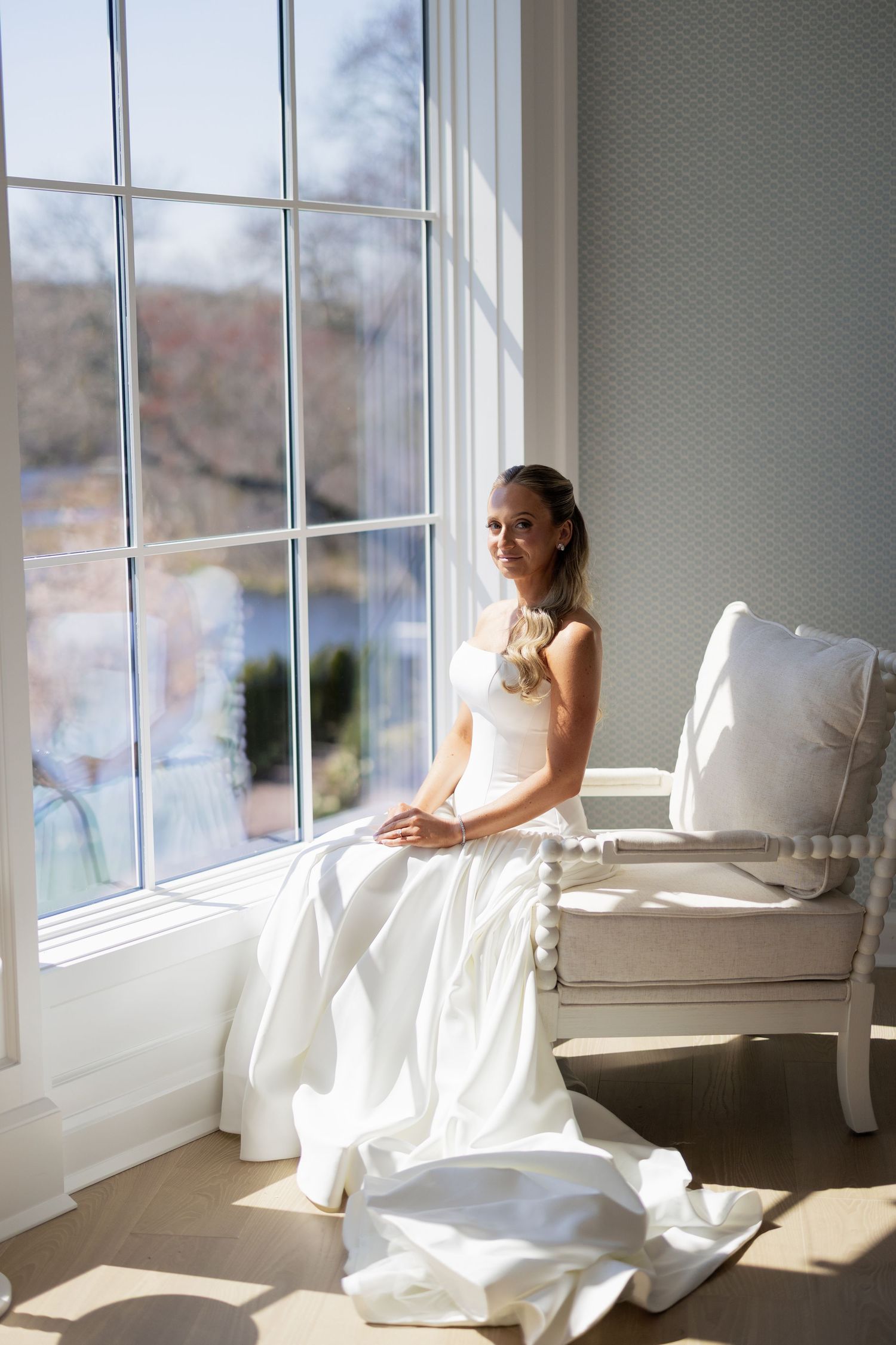 Bride in white gown sitting by sunlit window on elegant chair.