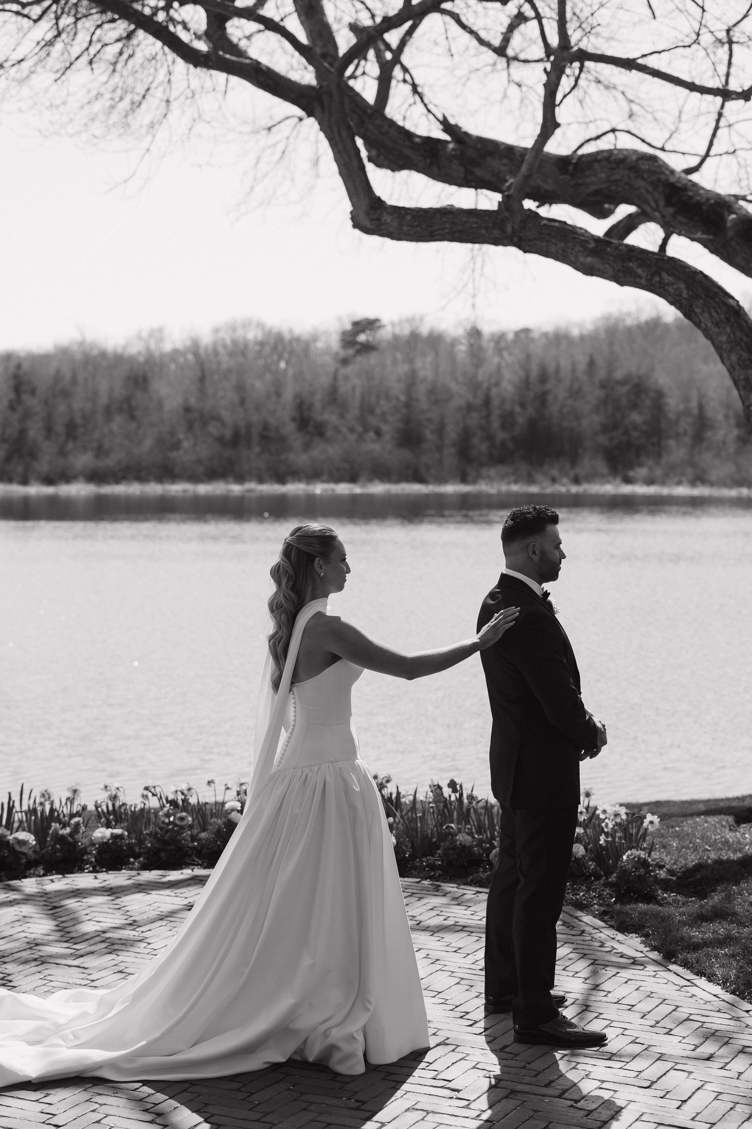 Black and white photo of a bride placing hands on groom's shoulders during a first look by a serene lakeside.