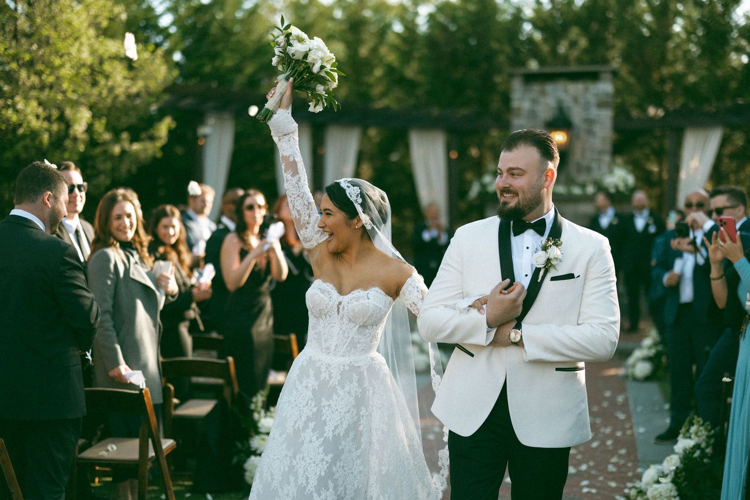 Wedding guests dancing and celebrating at an outdoor reception with trees and natural lighting.