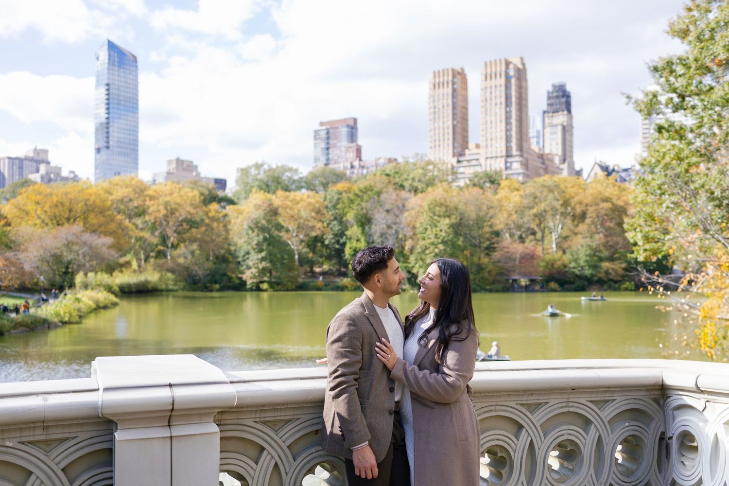 Panoramic view of Central Park lake with couple on stone bridge and city skyline behind.