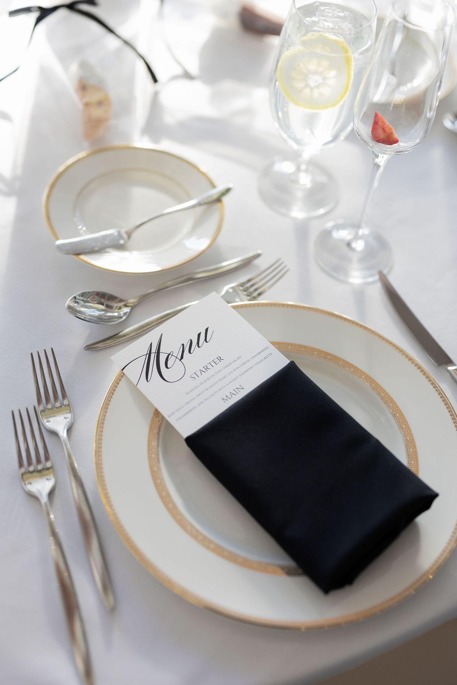Elegant wedding reception place setting with gold-rimmed plates, black napkin, menu card, and crystal glassware.