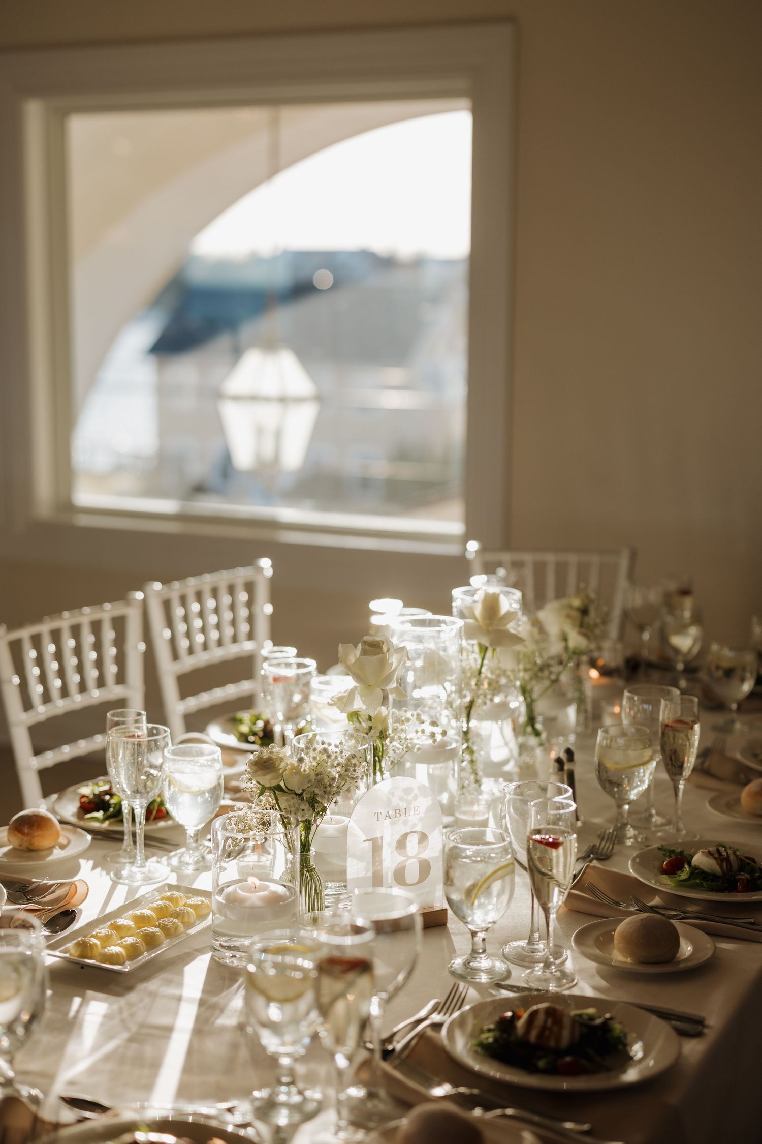 Elegant wedding reception tables set with white linens, glassware and floral centerpieces in natural window light.