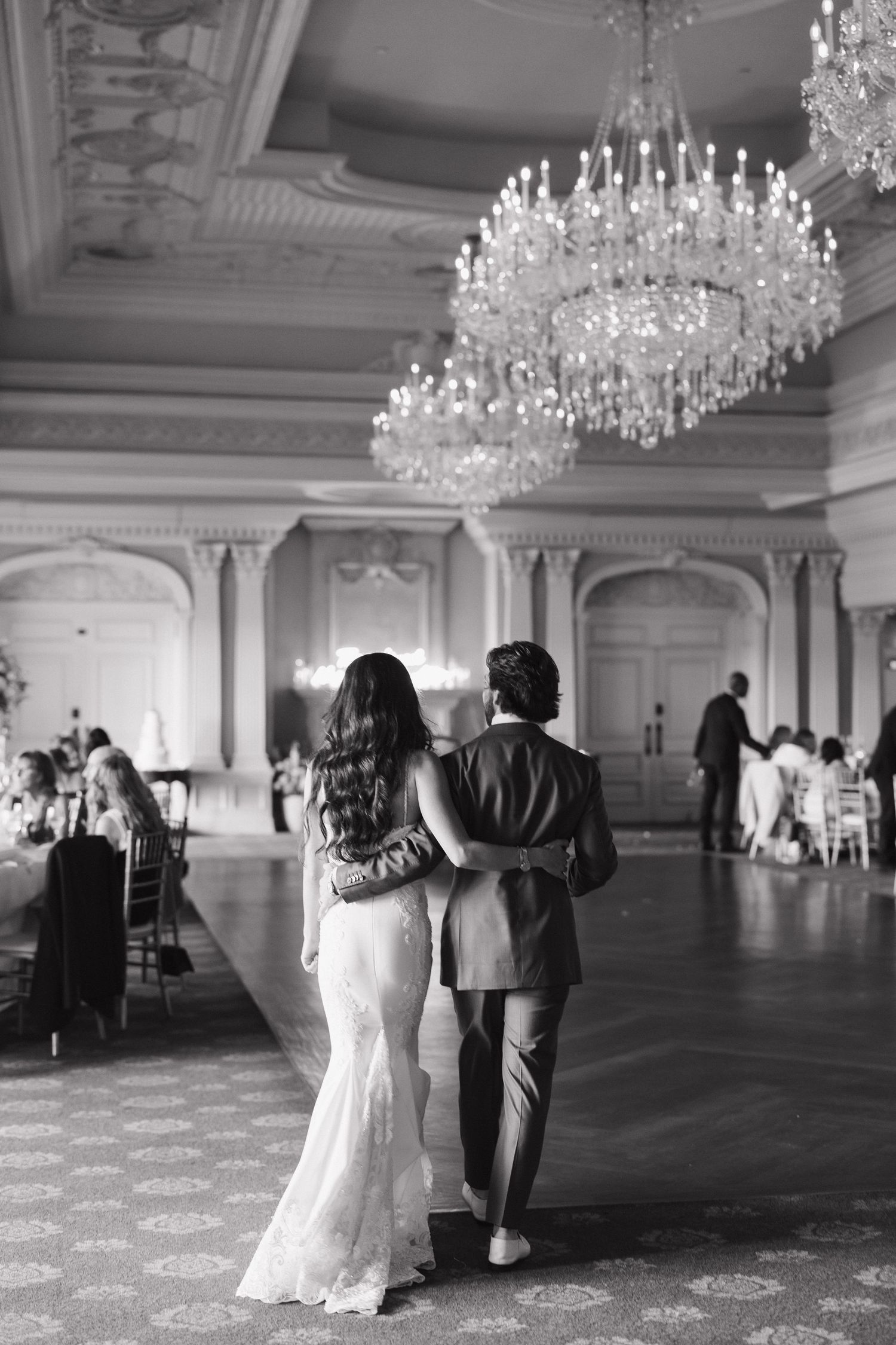 Black and white photo of couple walking in elegant ballroom under crystal chandeliers.