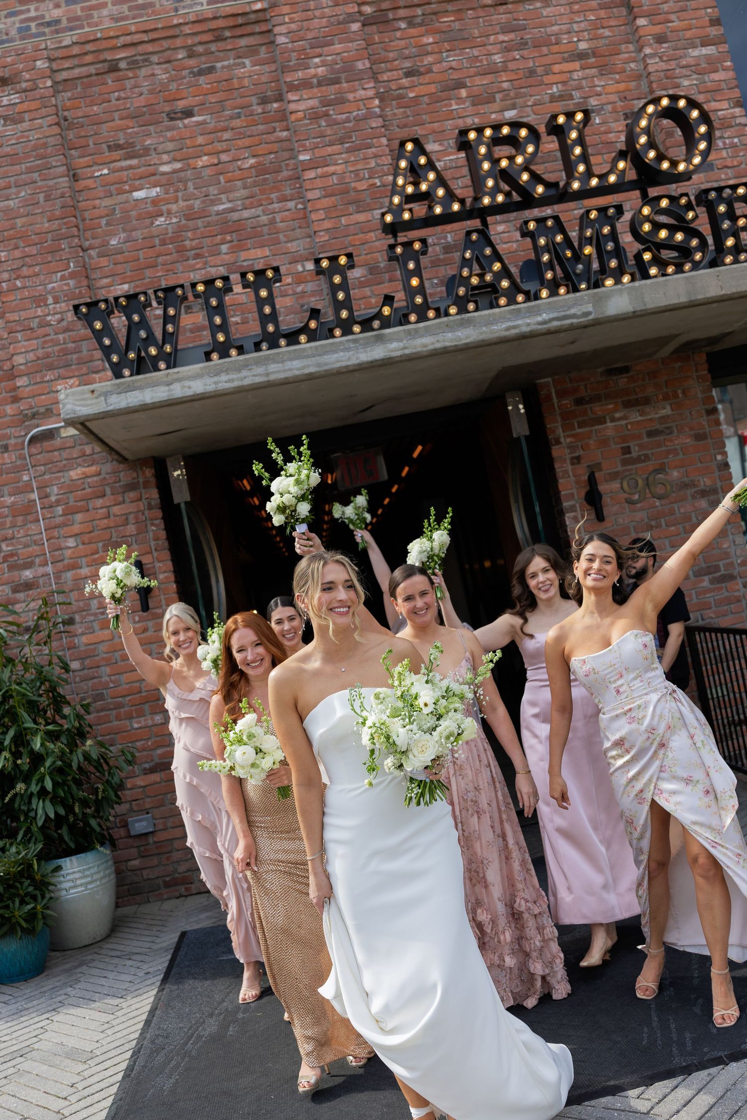 Wedding party celebrating and posing in front of a brick building with illuminated marquee signage.