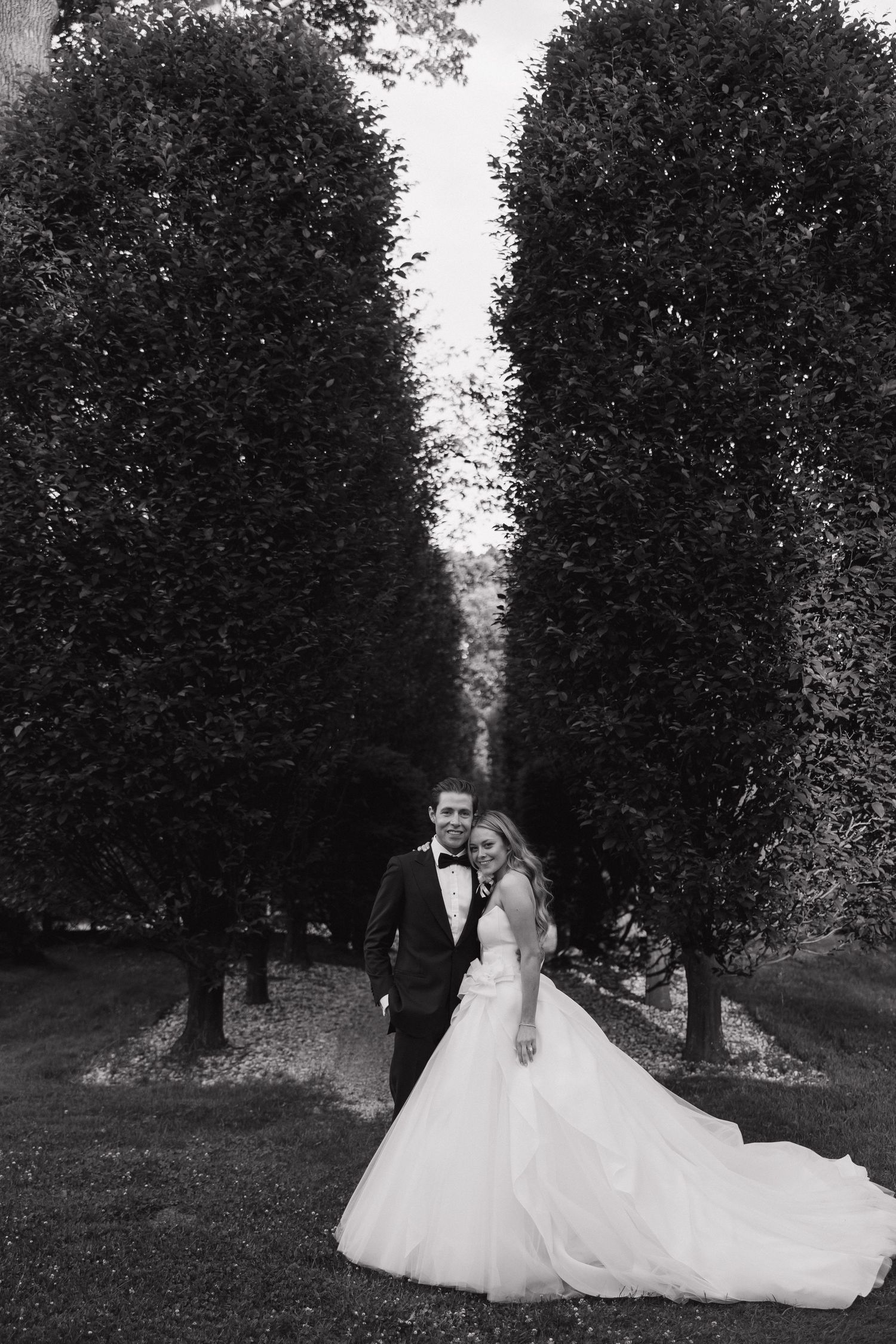 A dramatic black and white wedding portrait set between two tall hedges with a long dress train.