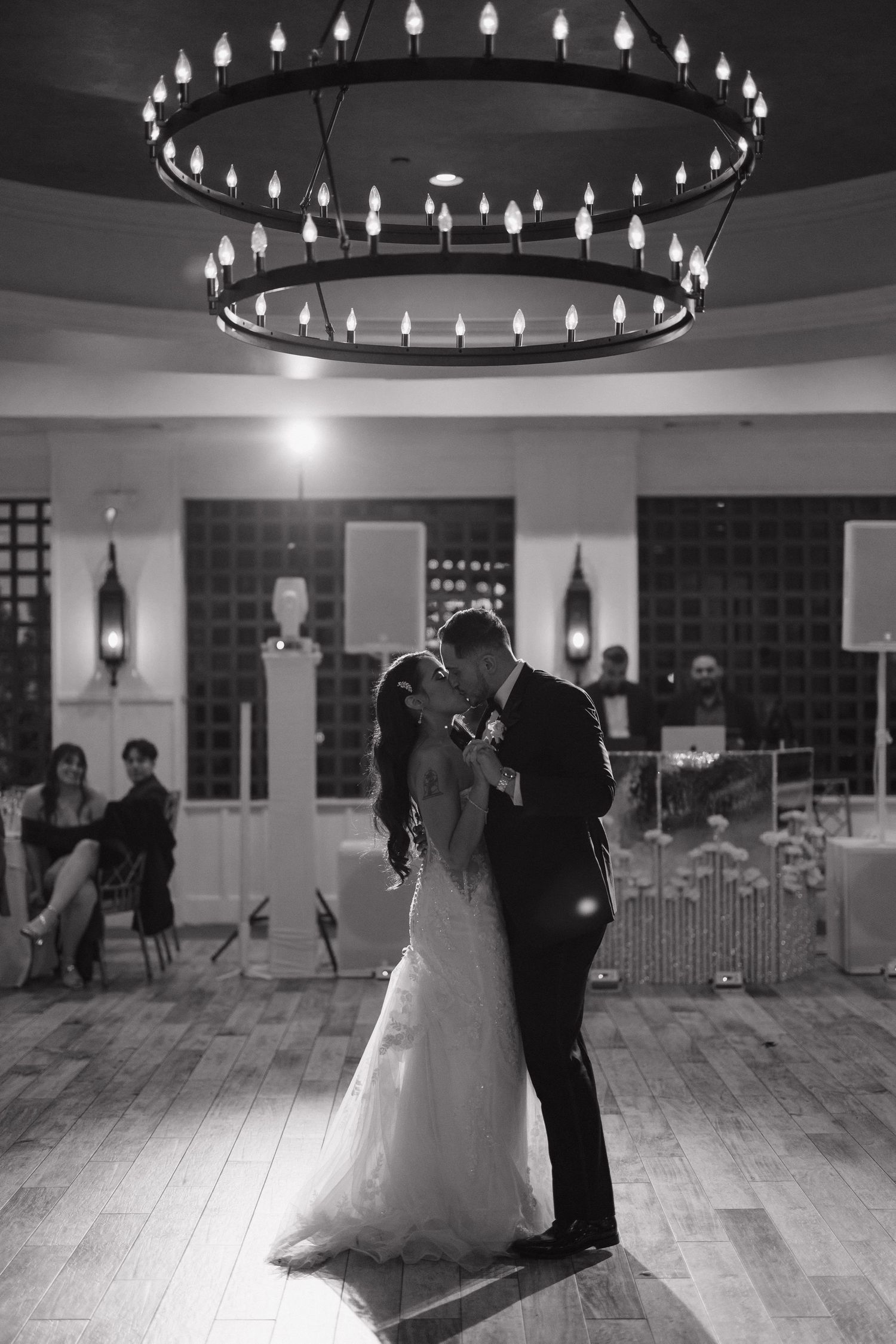 Black and white photo of a bride and groom sharing their first dance under an elegant chandelier at a wedding reception.