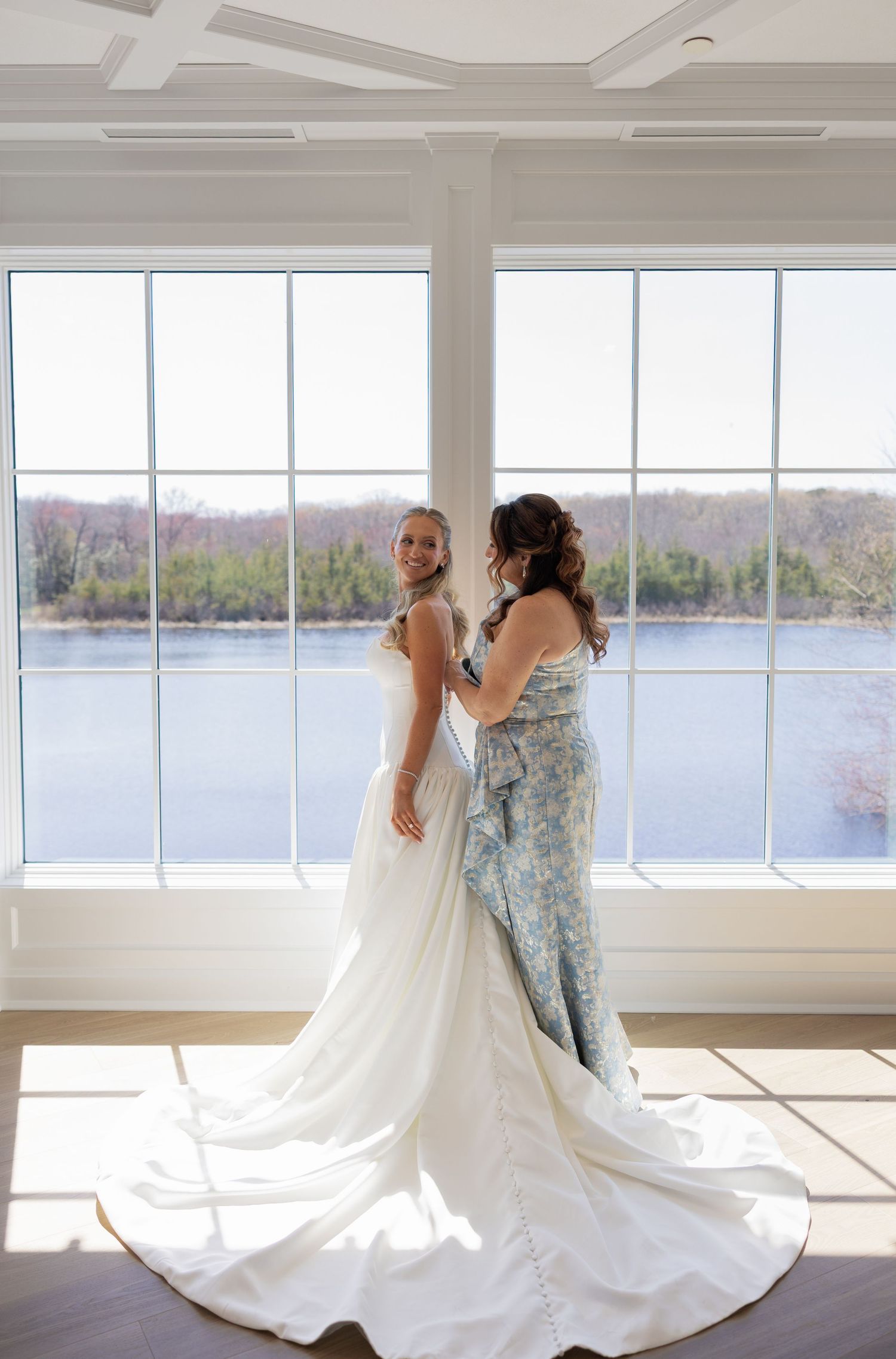 Bride and bridesmaid standing by large windows with river view.