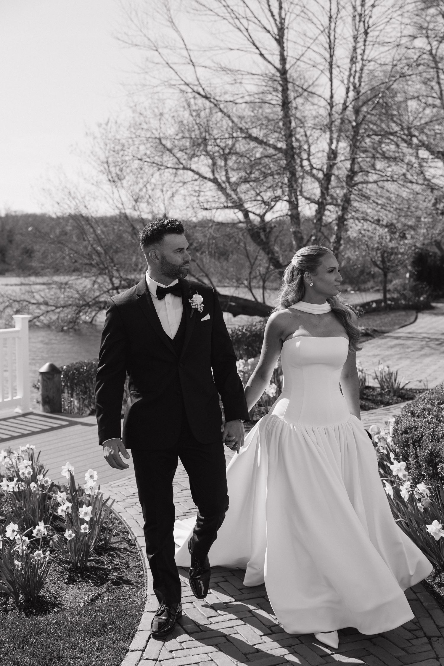 Black and white photo of bride and groom walking hand in hand through a garden on their wedding day.